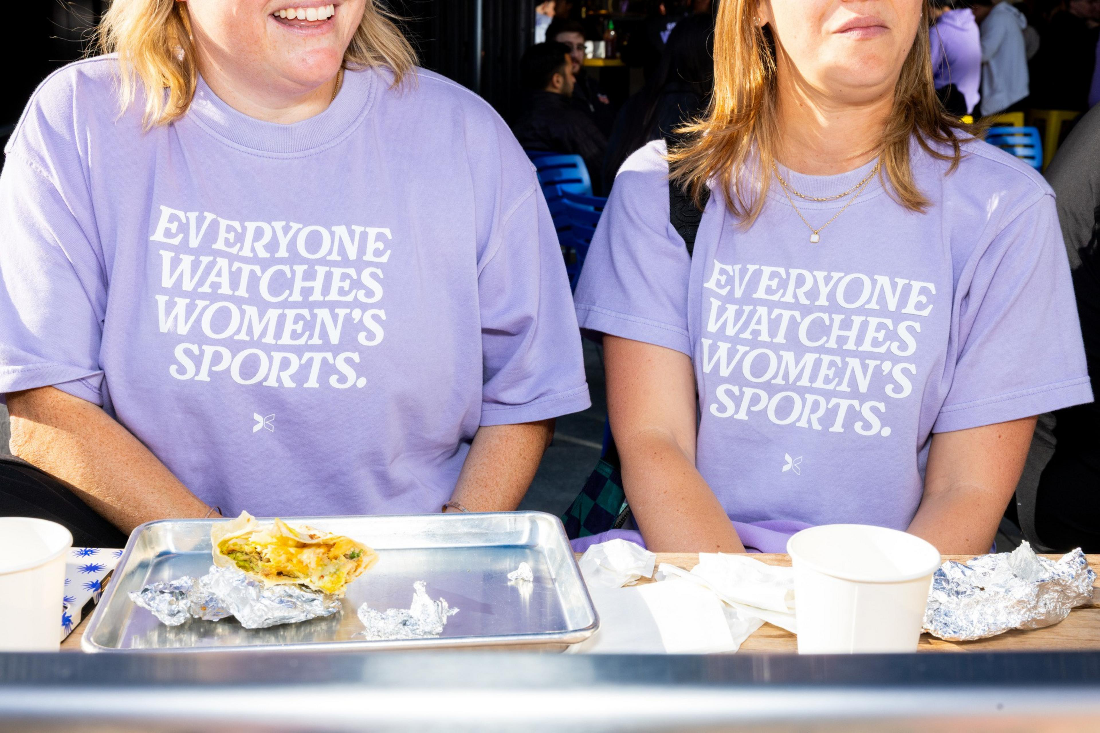 Two people wear matching lavender shirts with &quot;EVERYONE WATCHES WOMEN'S SPORTS&quot; printed on them. In front, there's a tray with food and a cup.