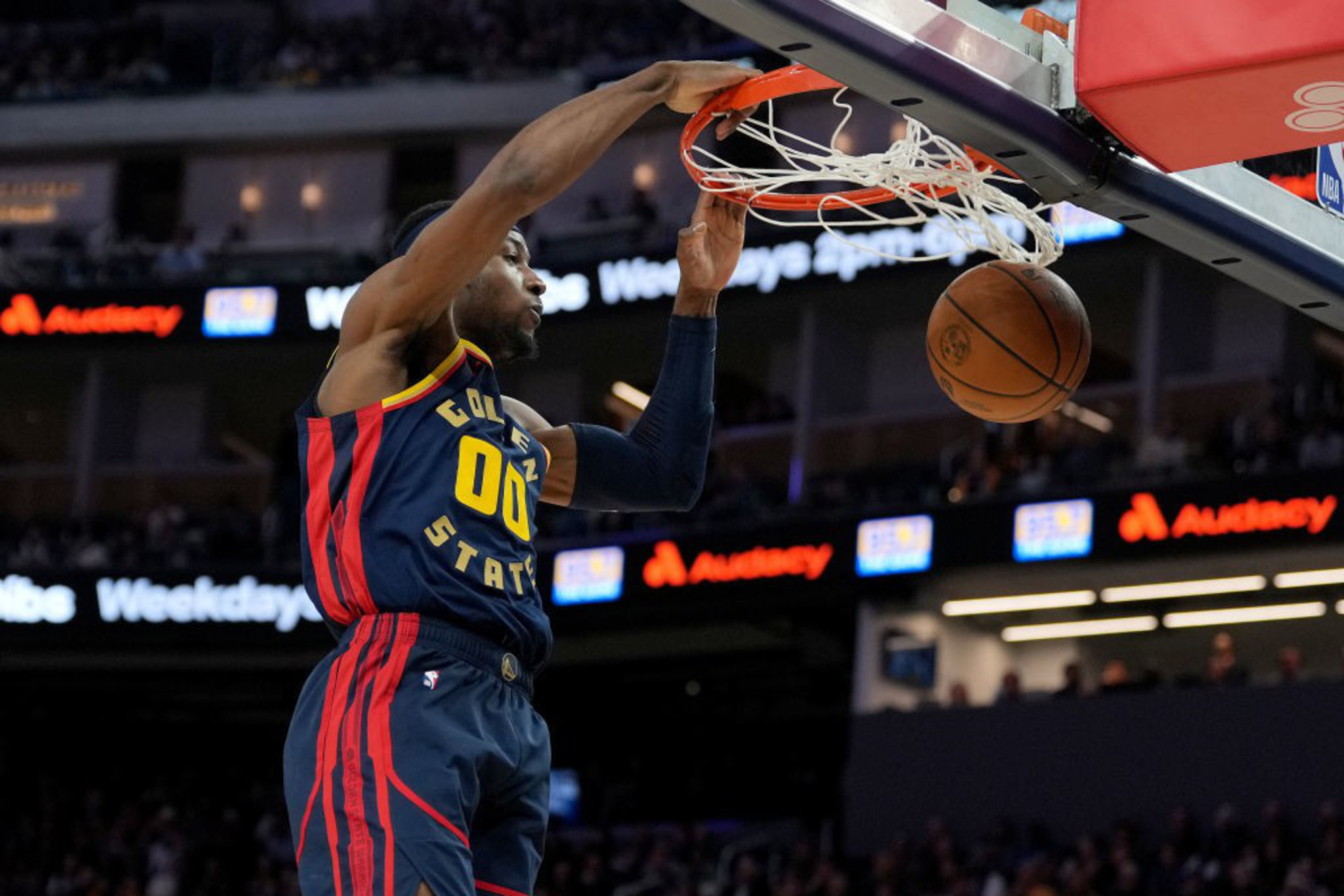A basketball player wearing a Golden State jersey is in mid-air about to dunk the ball into the hoop. The background shows a crowded arena.