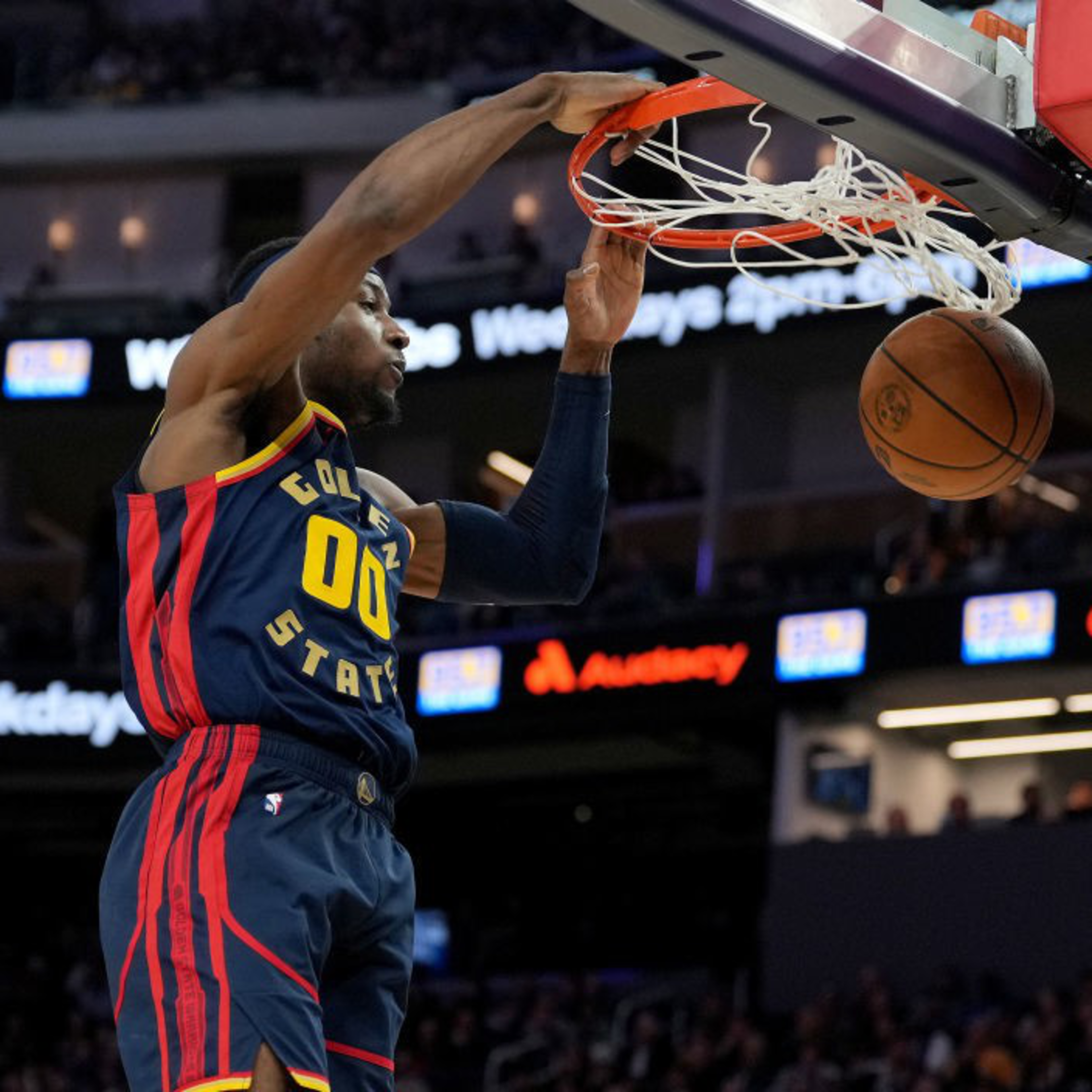 A basketball player wearing a Golden State jersey is in mid-air about to dunk the ball into the hoop. The background shows a crowded arena.