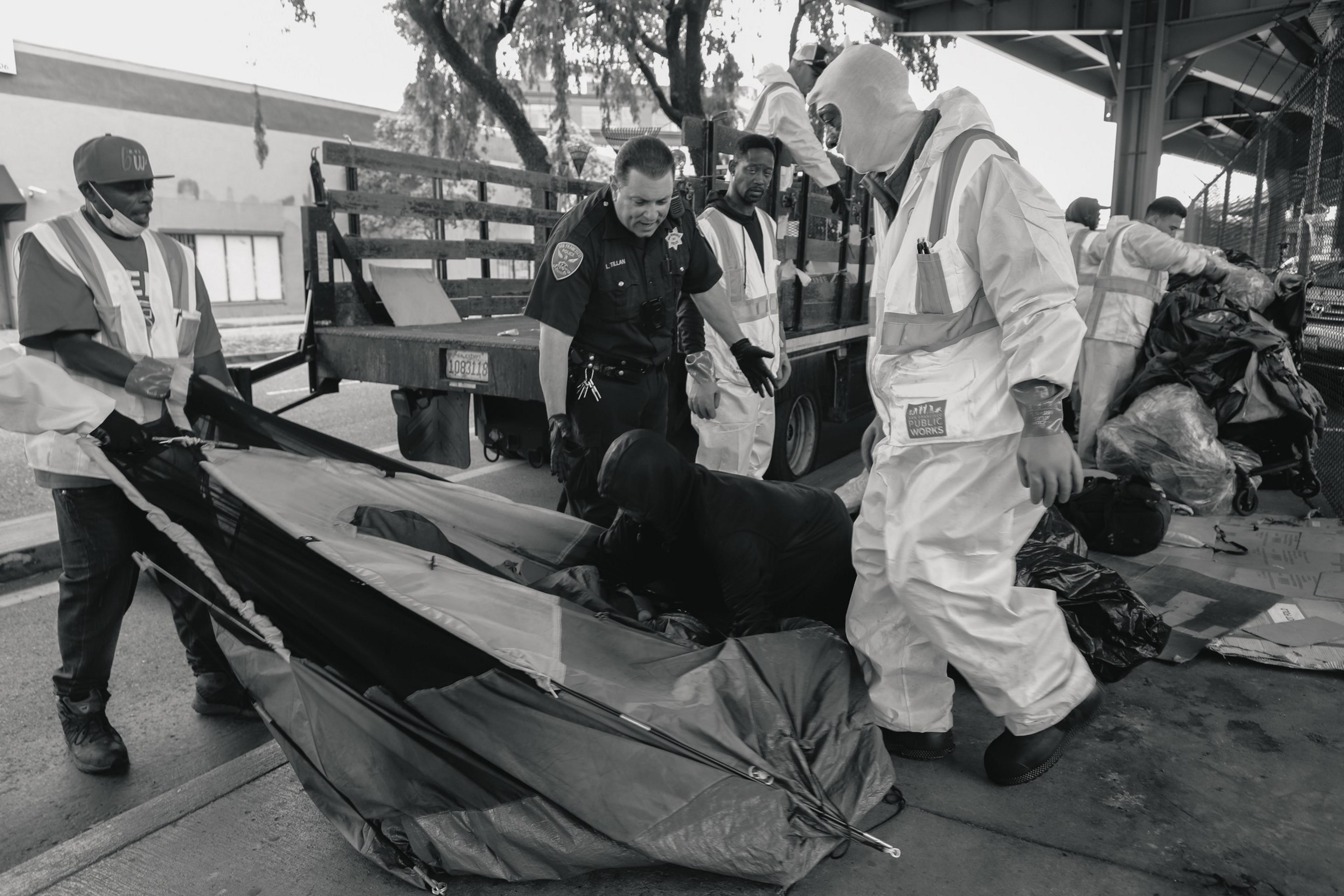 Several workers in safety gear and a police officer dismantle a tent under a shelter. One person, dressed in dark clothing, is inside the tent. A truck is in the background.