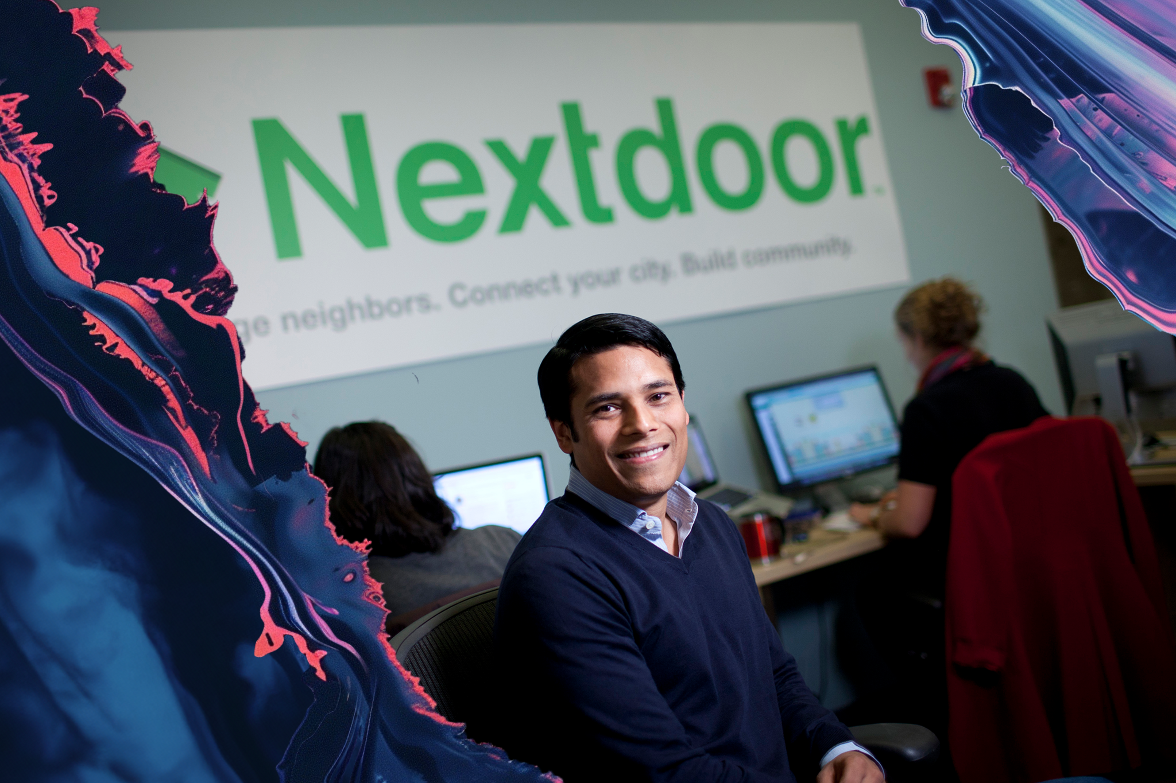 A man is smiling and sitting in an office with two others working on computers. The word "Nextdoor" is prominently displayed on a wall behind them.