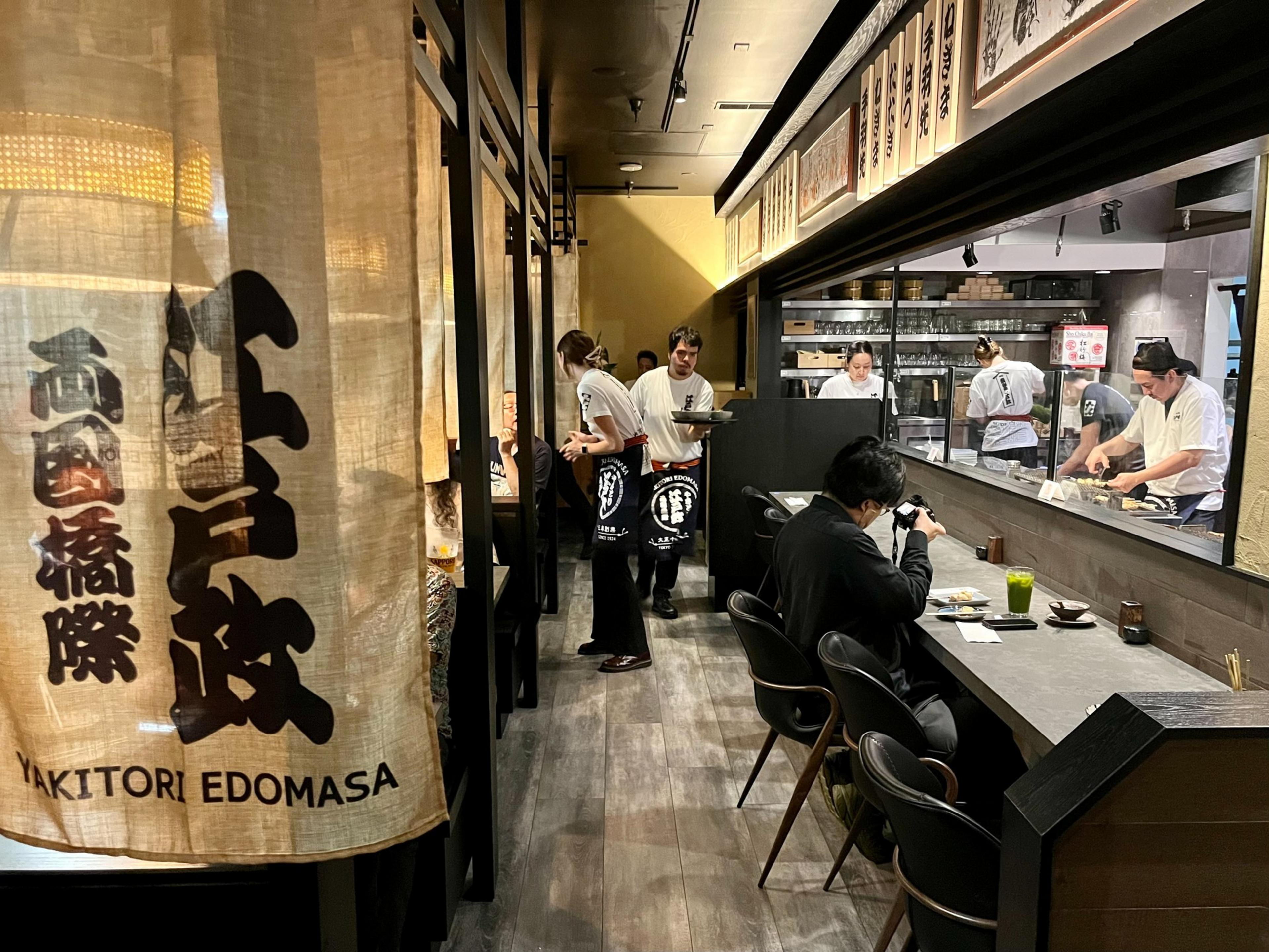 A Japanese restaurant with a fabric curtain, diners seated at a counter and tables, waitstaff serving, and chefs preparing food behind a glass window.