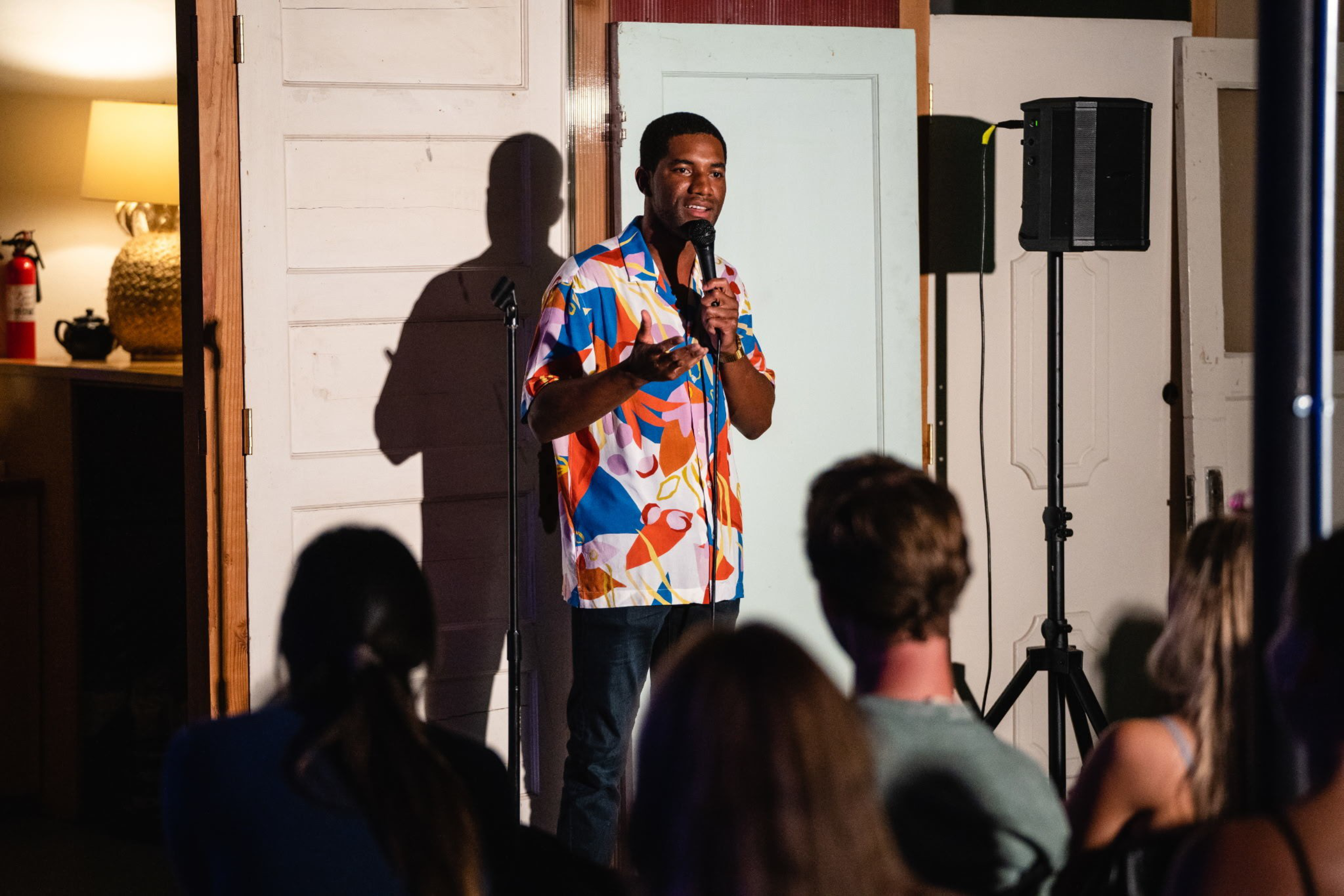 A man in a colorful shirt stands on stage holding a microphone, speaking to an audience with a speaker and vintage doors in the background.