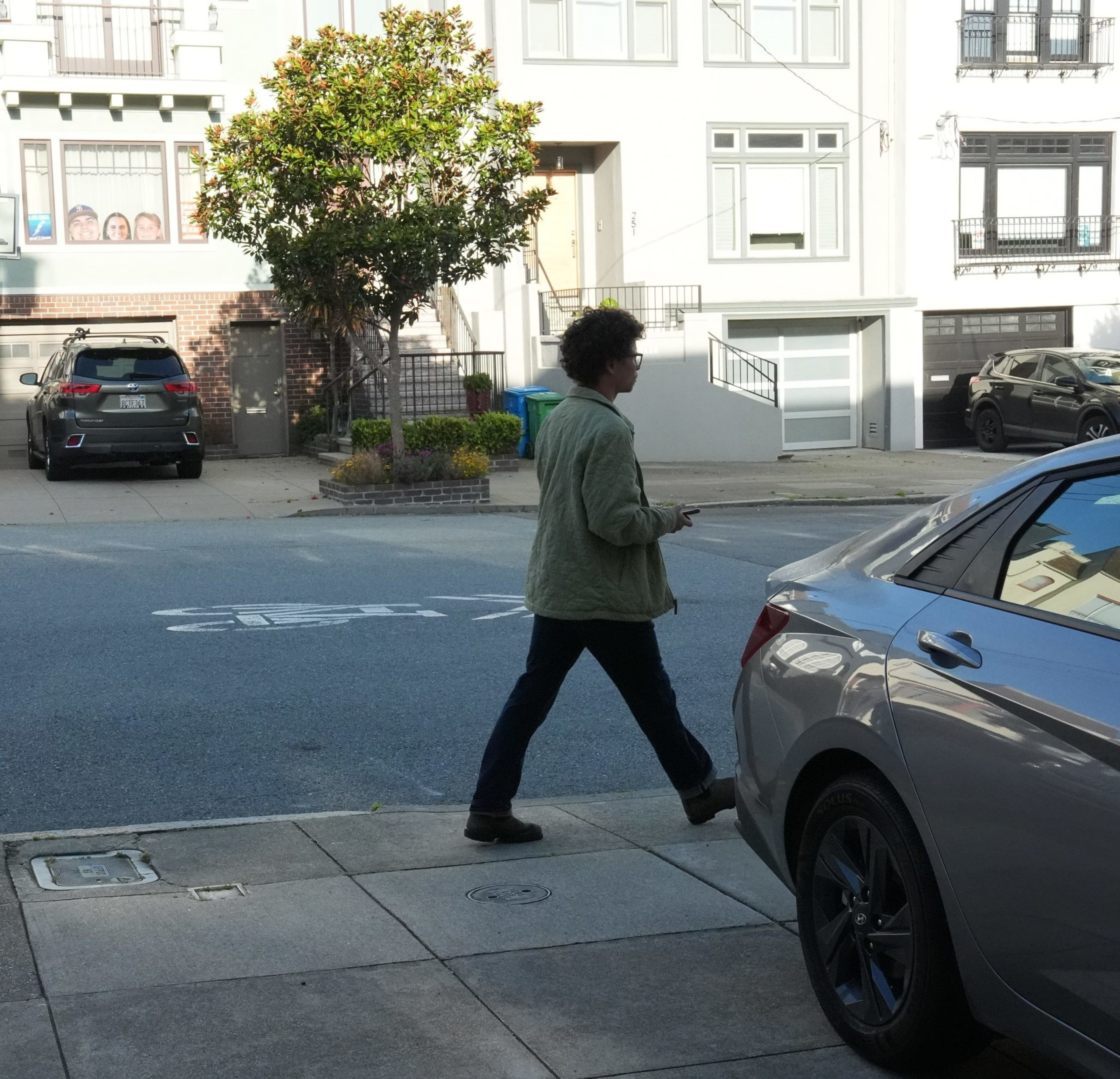 A person walks past parked cars on a sunny residential street with houses and a basketball hoop.
