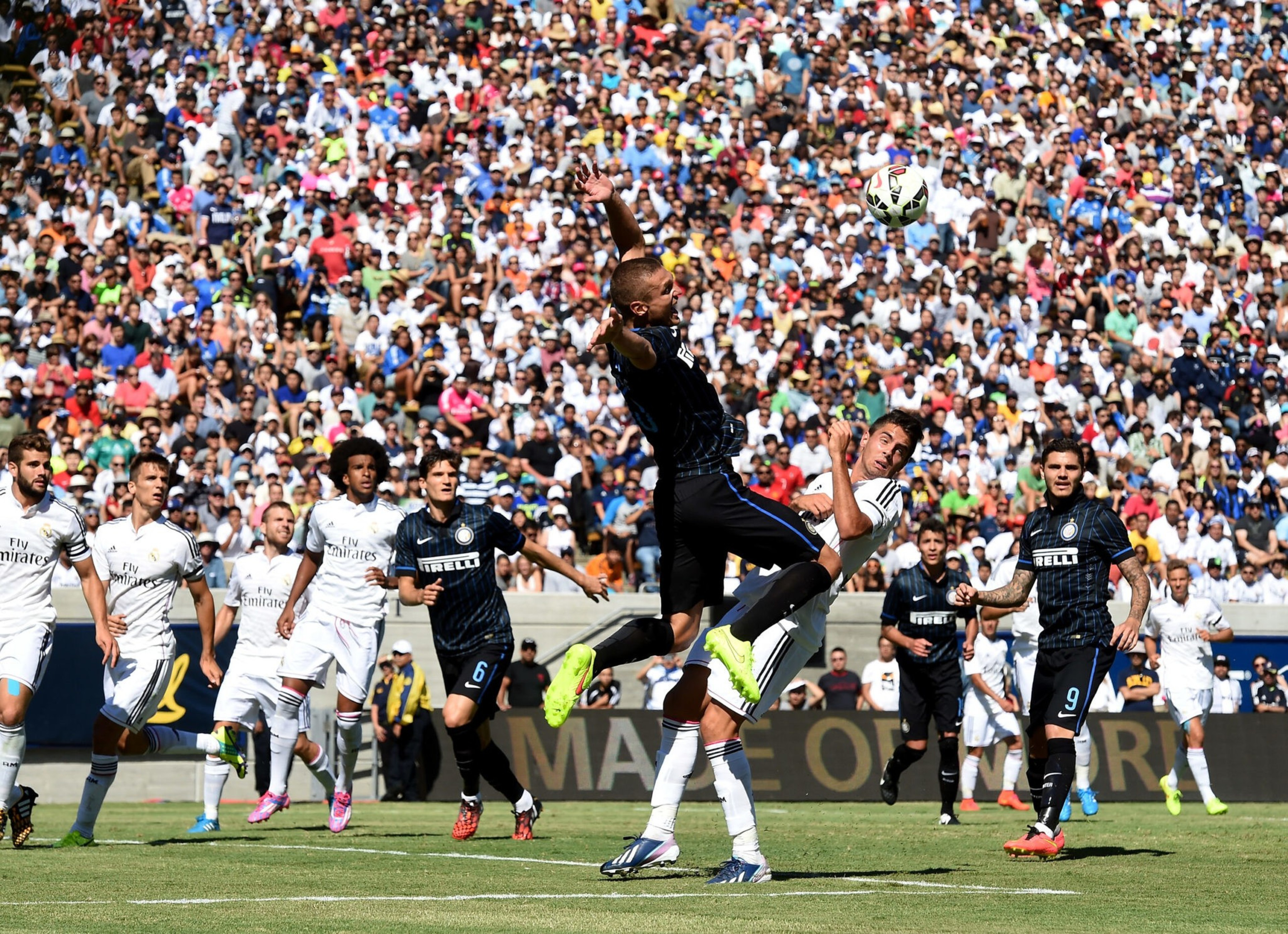 Two soccer players jump to head the ball mid-air, surrounded by teammates and opponents, with a large crowd watching from the stands.