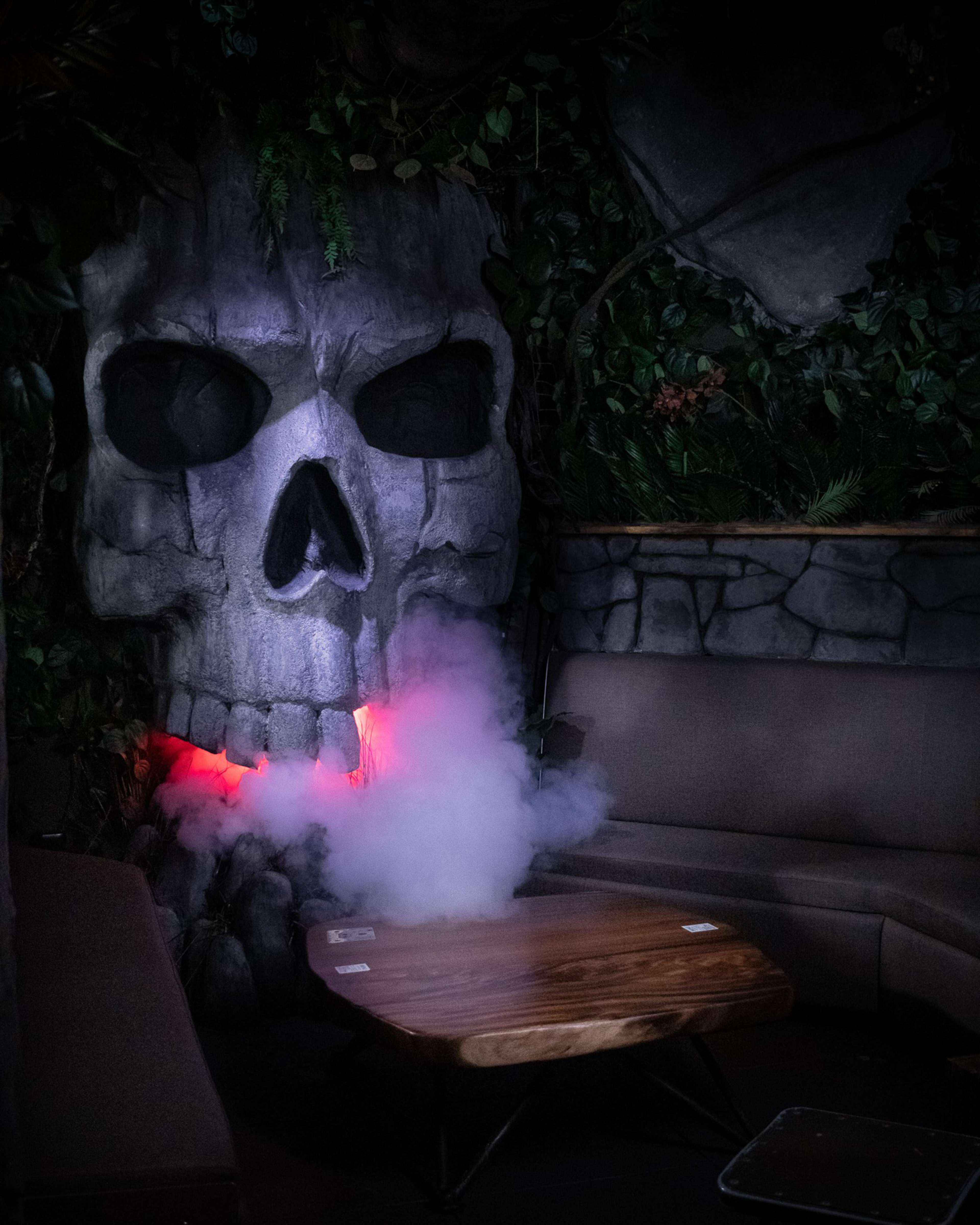A large stone skull with glowing red light and mist emanating from its mouth is set beside a wooden table and brown seating against a leafy backdrop.