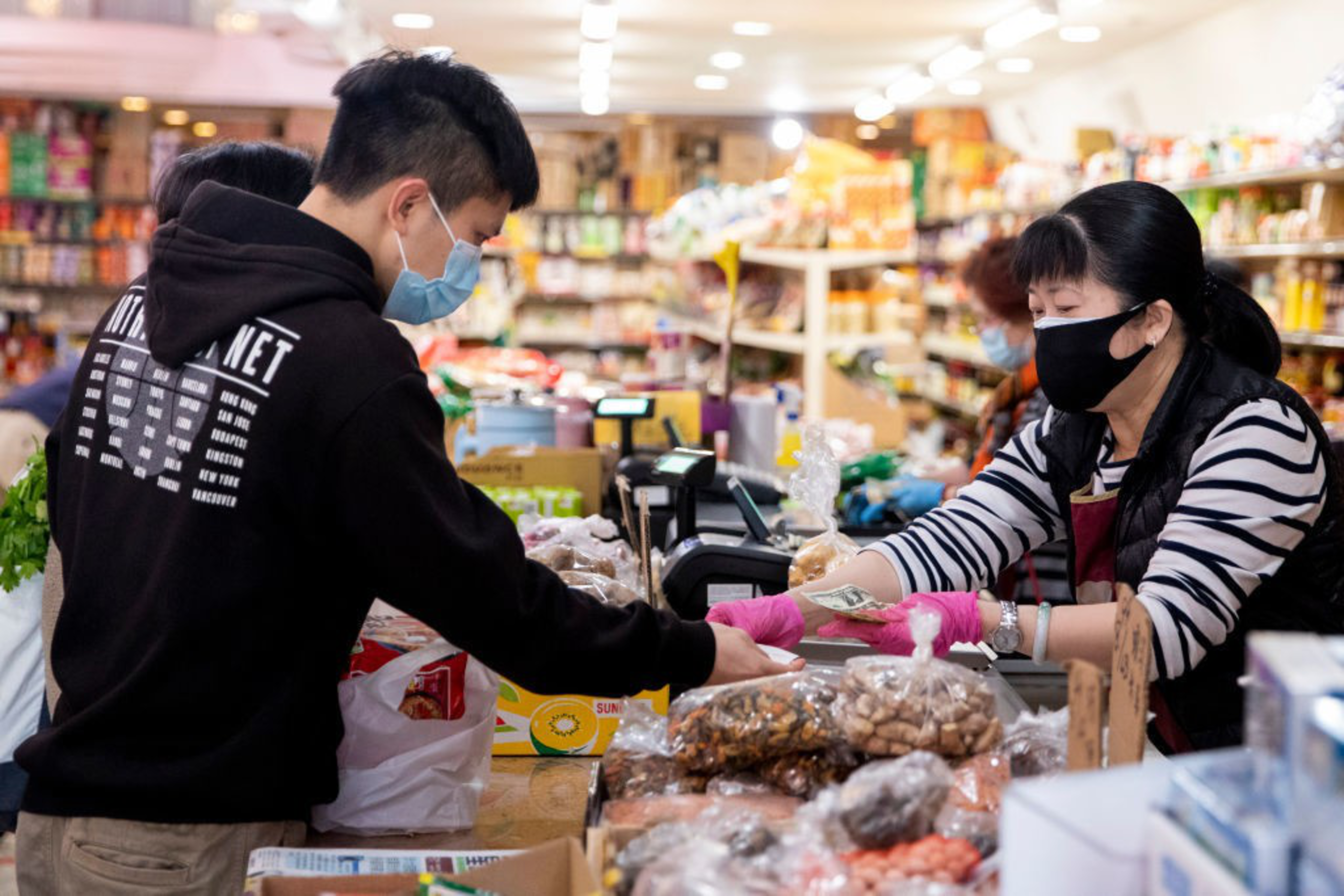 A person in a mask is handing cash to a masked cashier at a grocery store checkout counter.