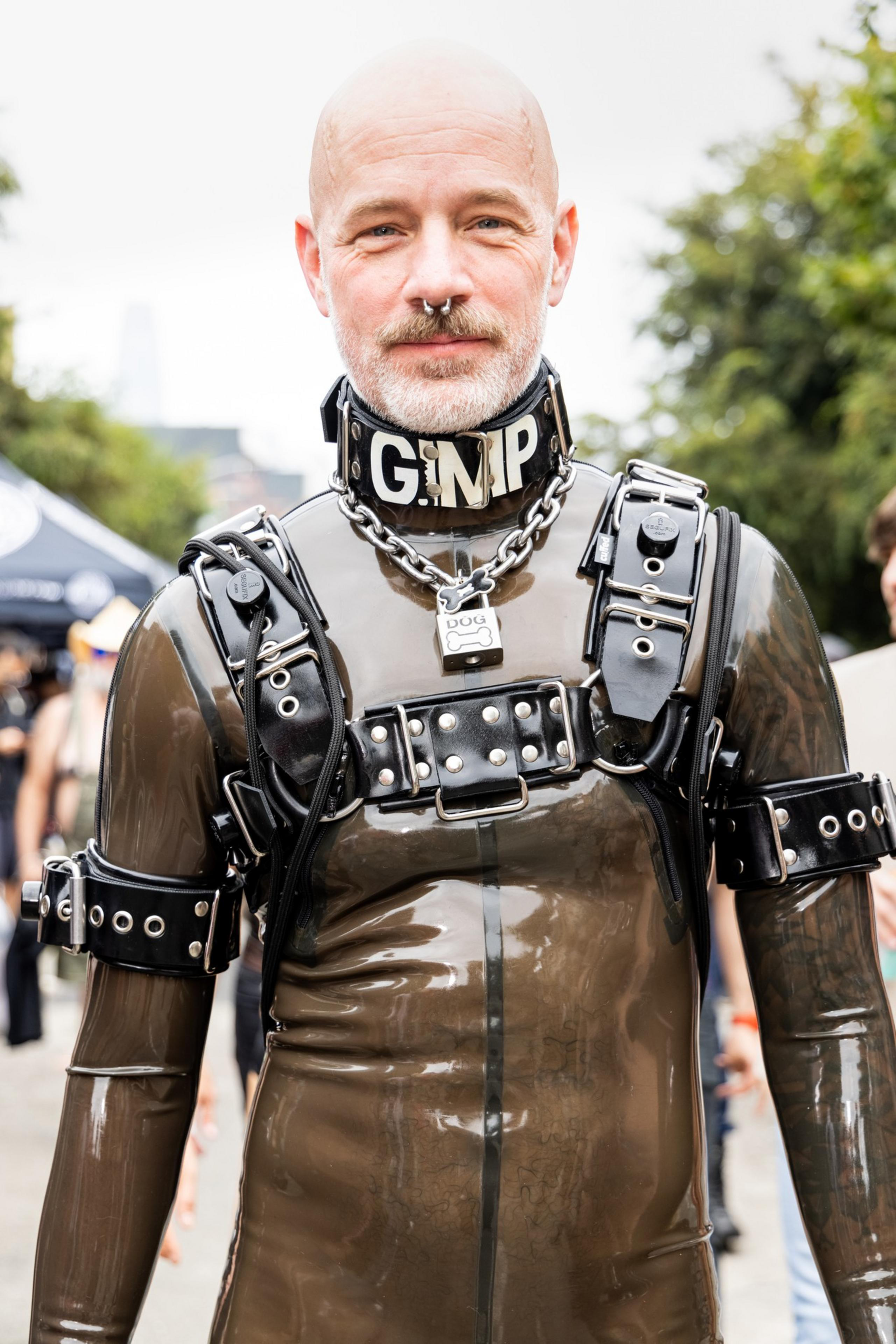 A bald man wears a tight, shiny black latex suit with leather harnesses, a collar reading “GIMP,” a chain, and a padlock labeled “DOG.”