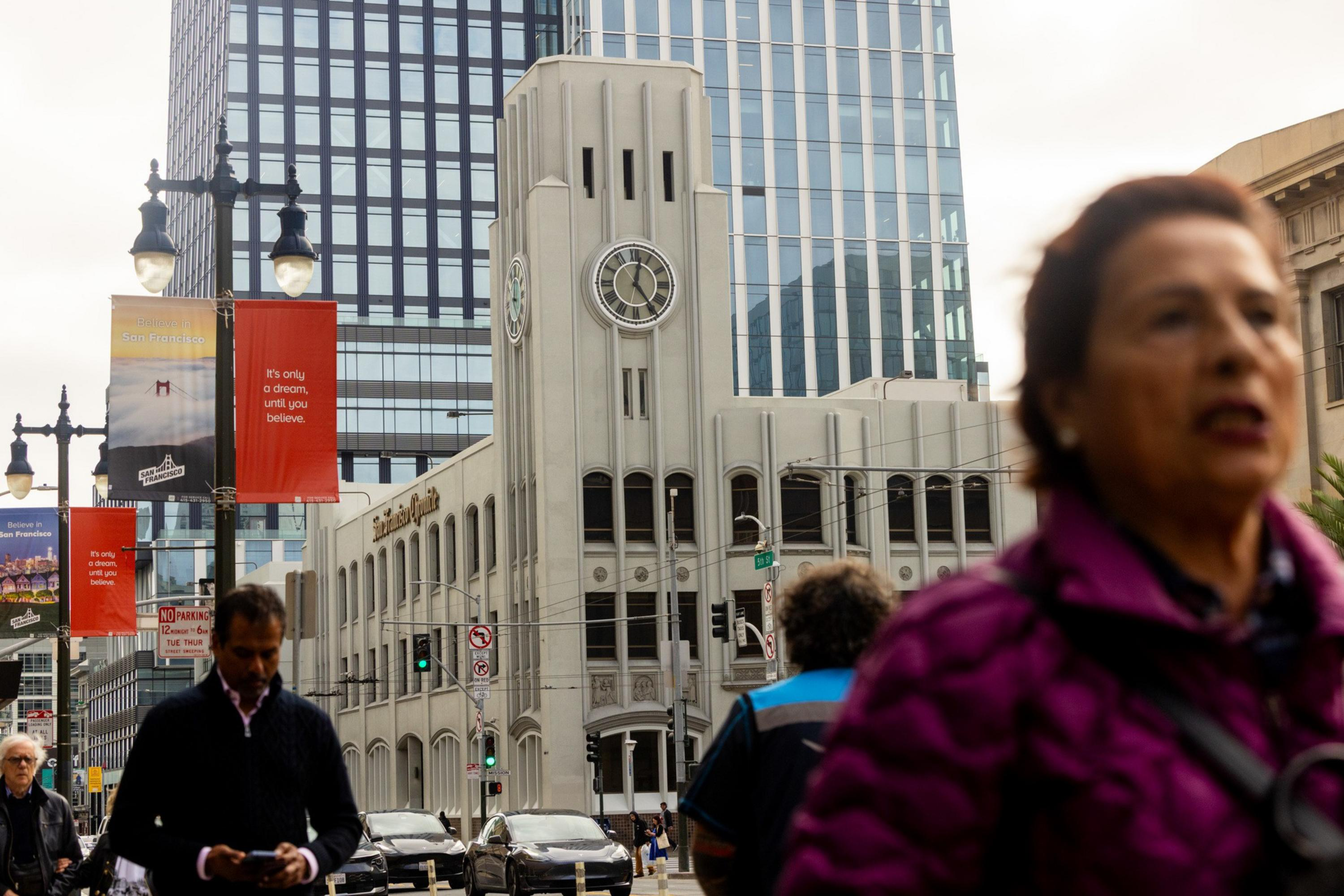 A white clock tower building stands against a modern glass skyscraper with people walking on the busy city street below.