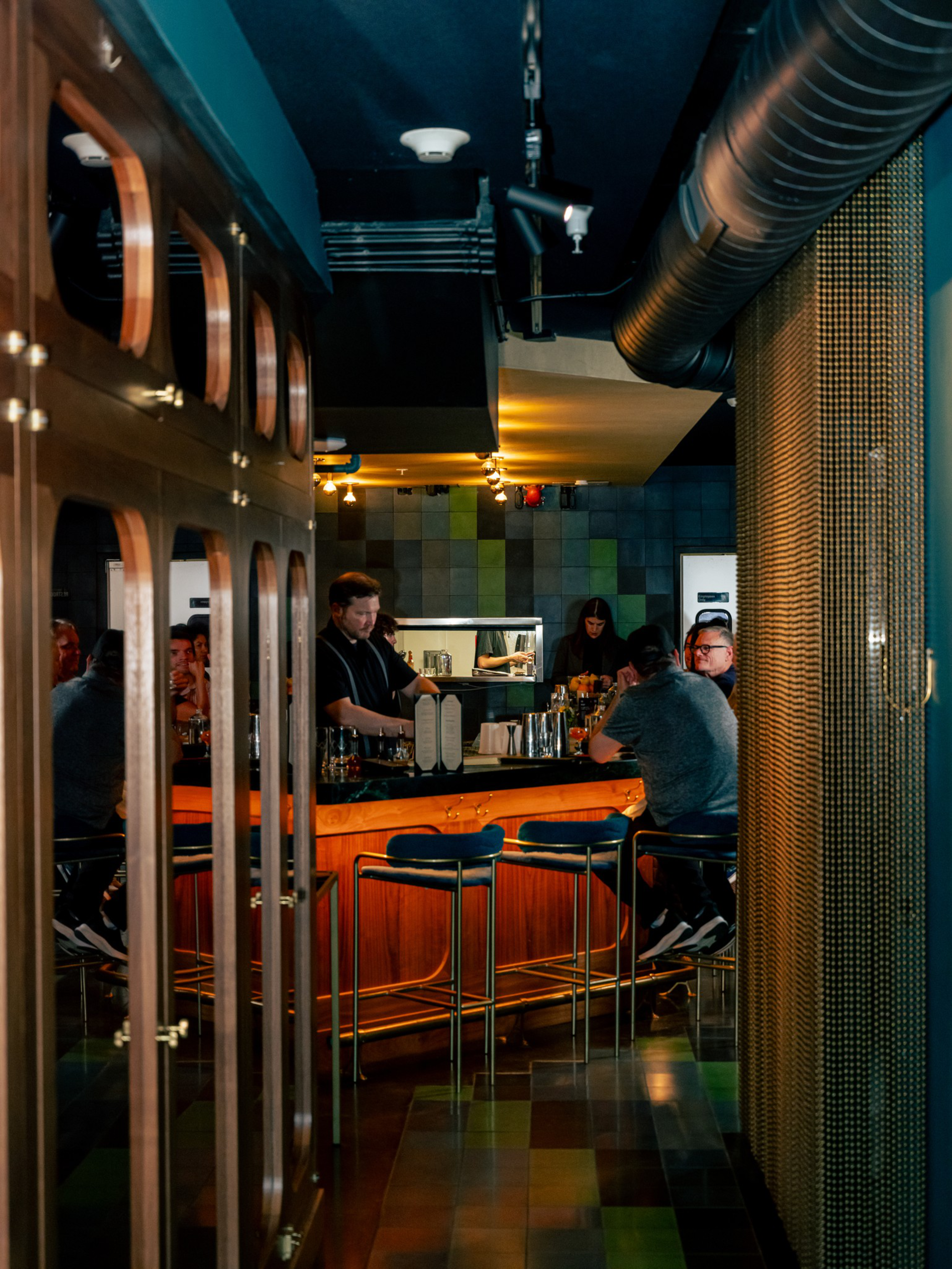 A dimly lit bar with a wooden counter, three blue bar stools, and several people seated and talking, with a bartender behind the counter.