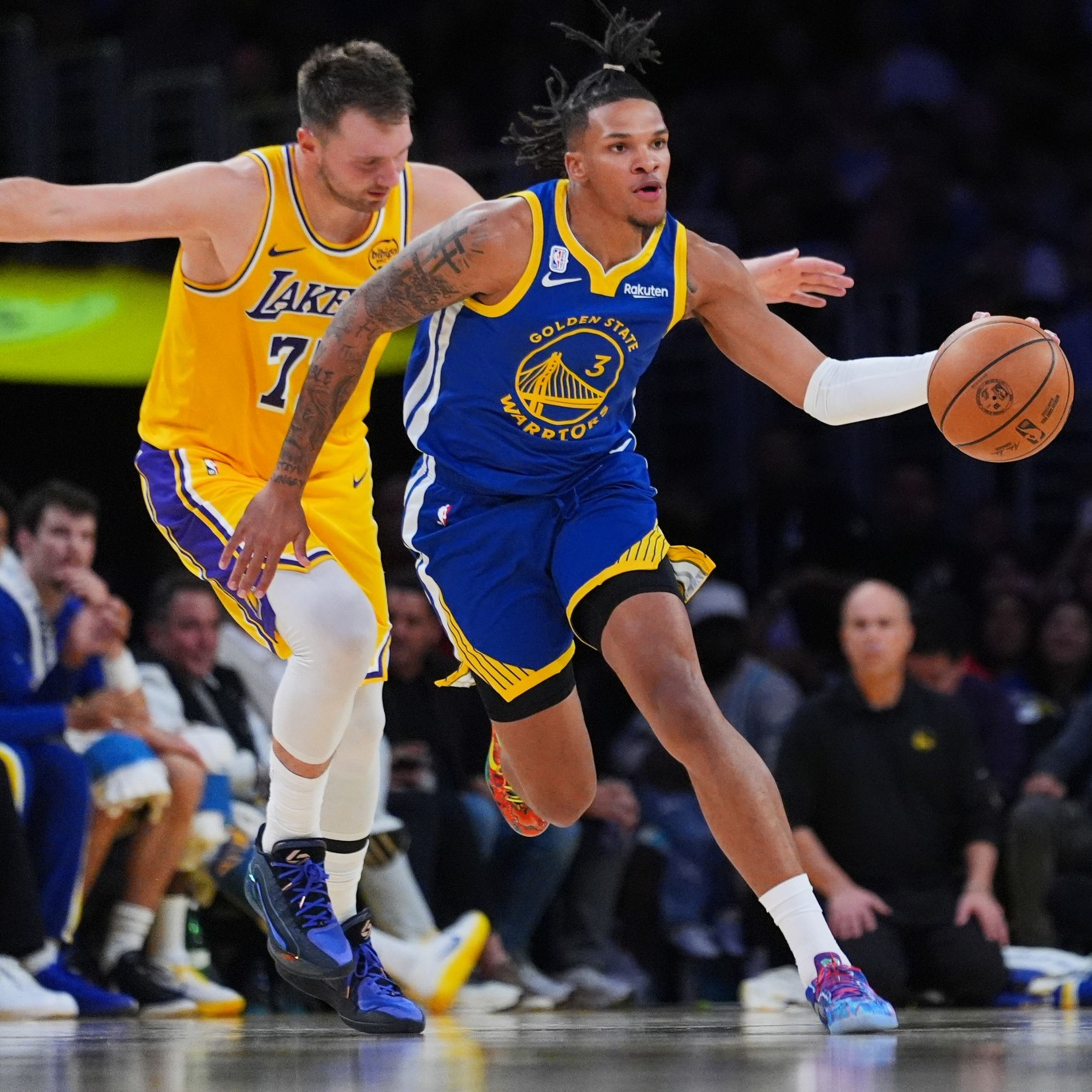 A Golden State Warriors player dribbles the basketball past a Los Angeles Lakers defender during a game, with spectators visible in the background.