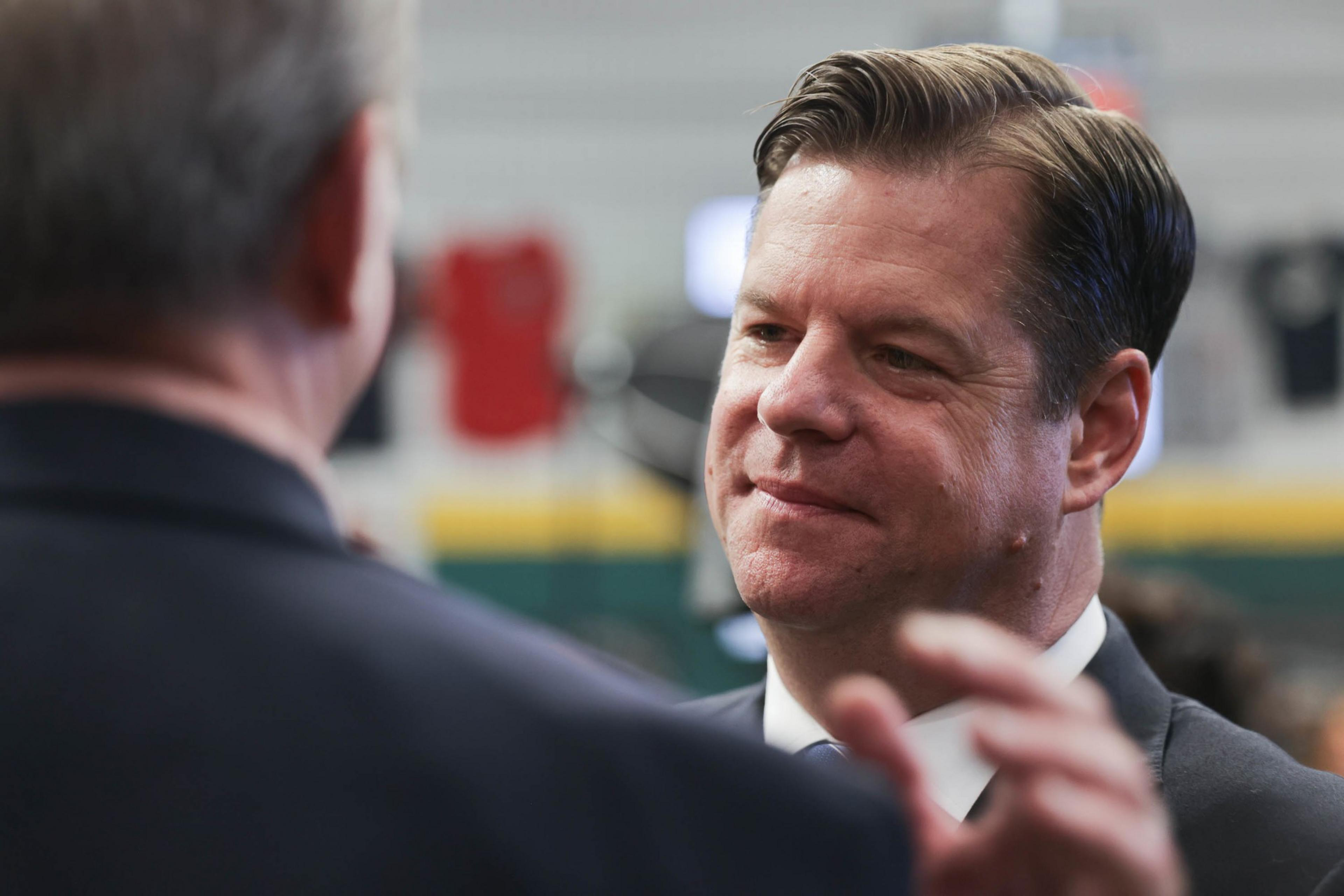 A man with slicked-back hair listens attentively to someone out of frame, in a blurred-industrial-setting background.
