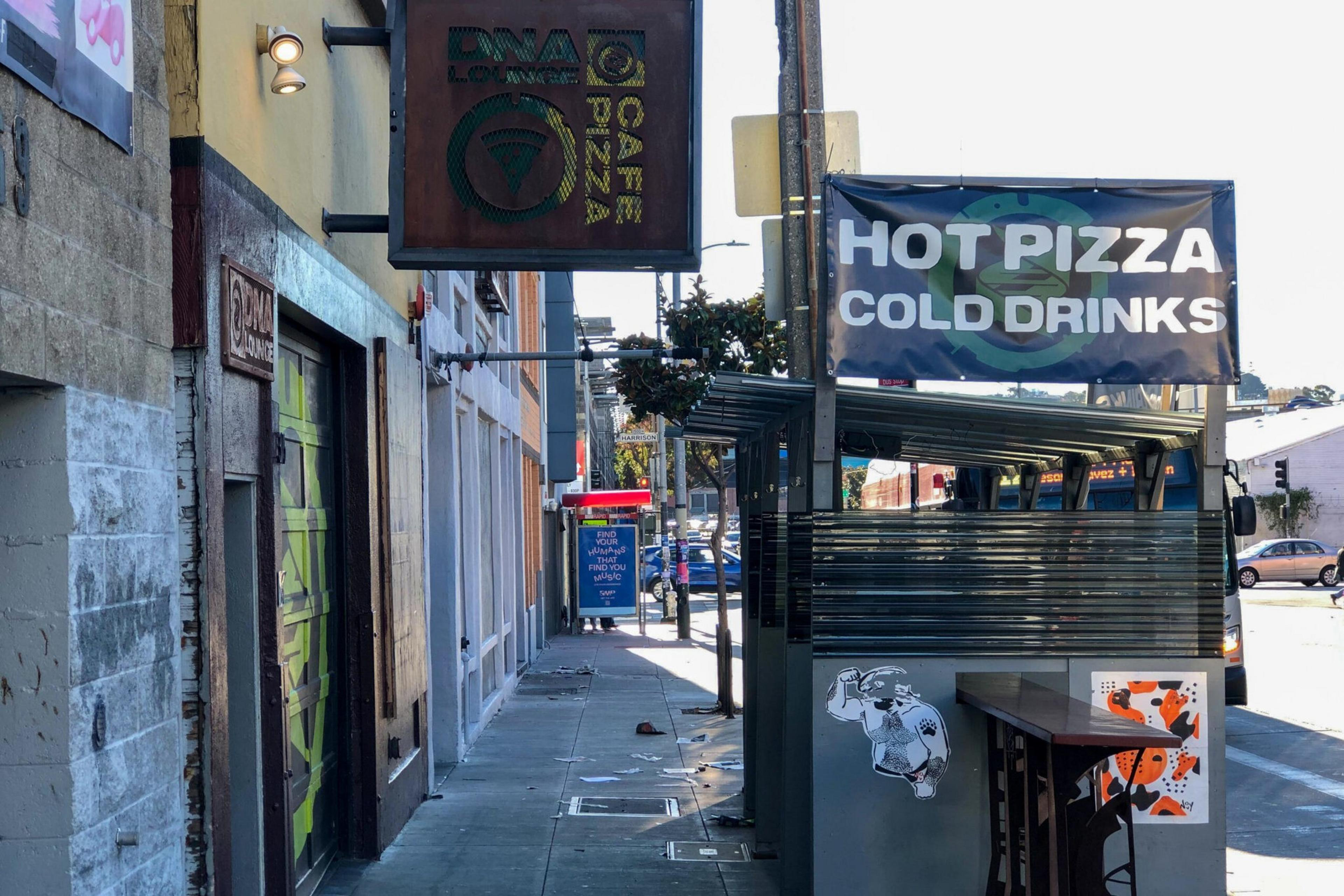 A street view of a storefront with signs advertising "DNA Lounge Cafe Pizza" and a banner for "Hot Pizza Cold Drinks." A small outdoor seating area is visible.