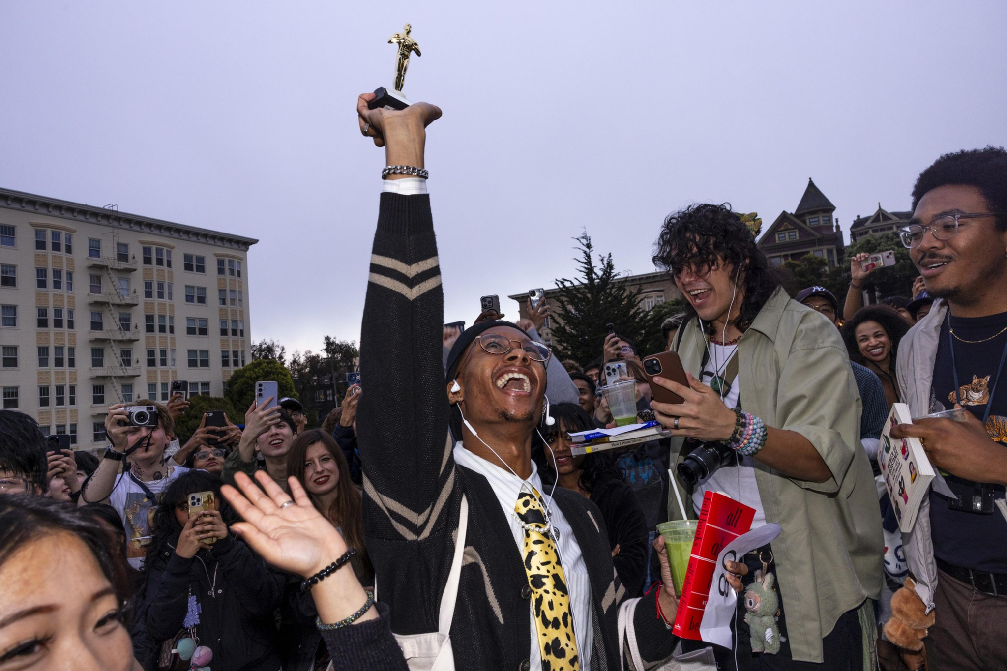 A joyful person in a leopard-print tie raises a trophy high while surrounded by an excited crowd taking photos and celebrating.