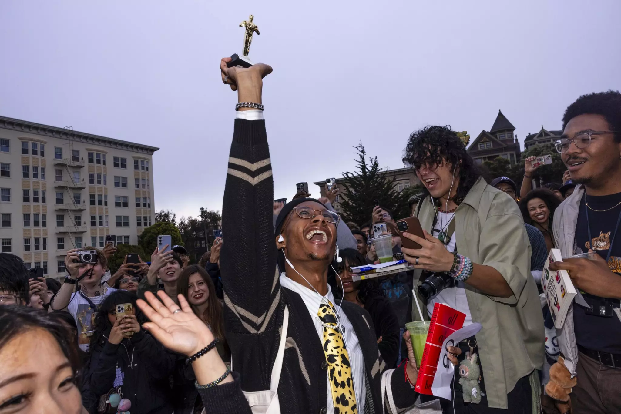 A joyful person in a leopard-print tie raises a trophy high while surrounded by an excited crowd taking photos and celebrating.