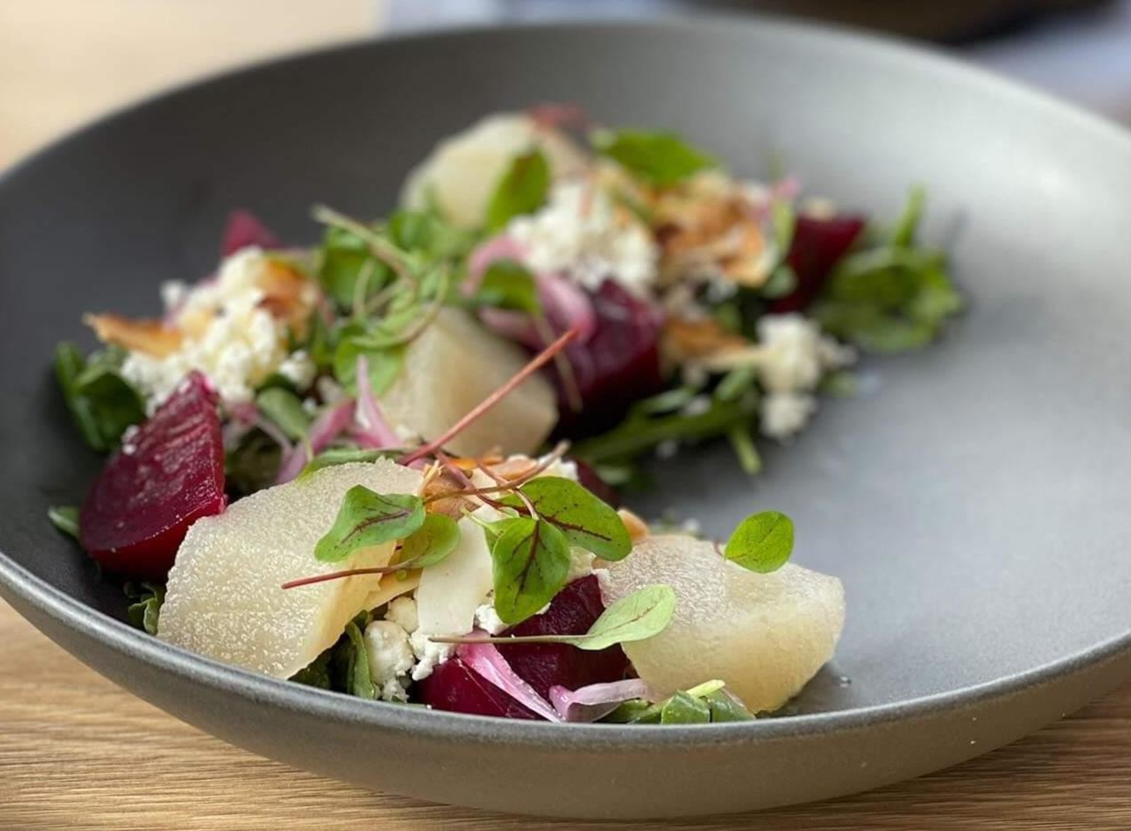 Slices of pear, beetroot, feta cheese, microgreens, and red onion are arranged on a dark plate, creating a fresh and colorful salad.