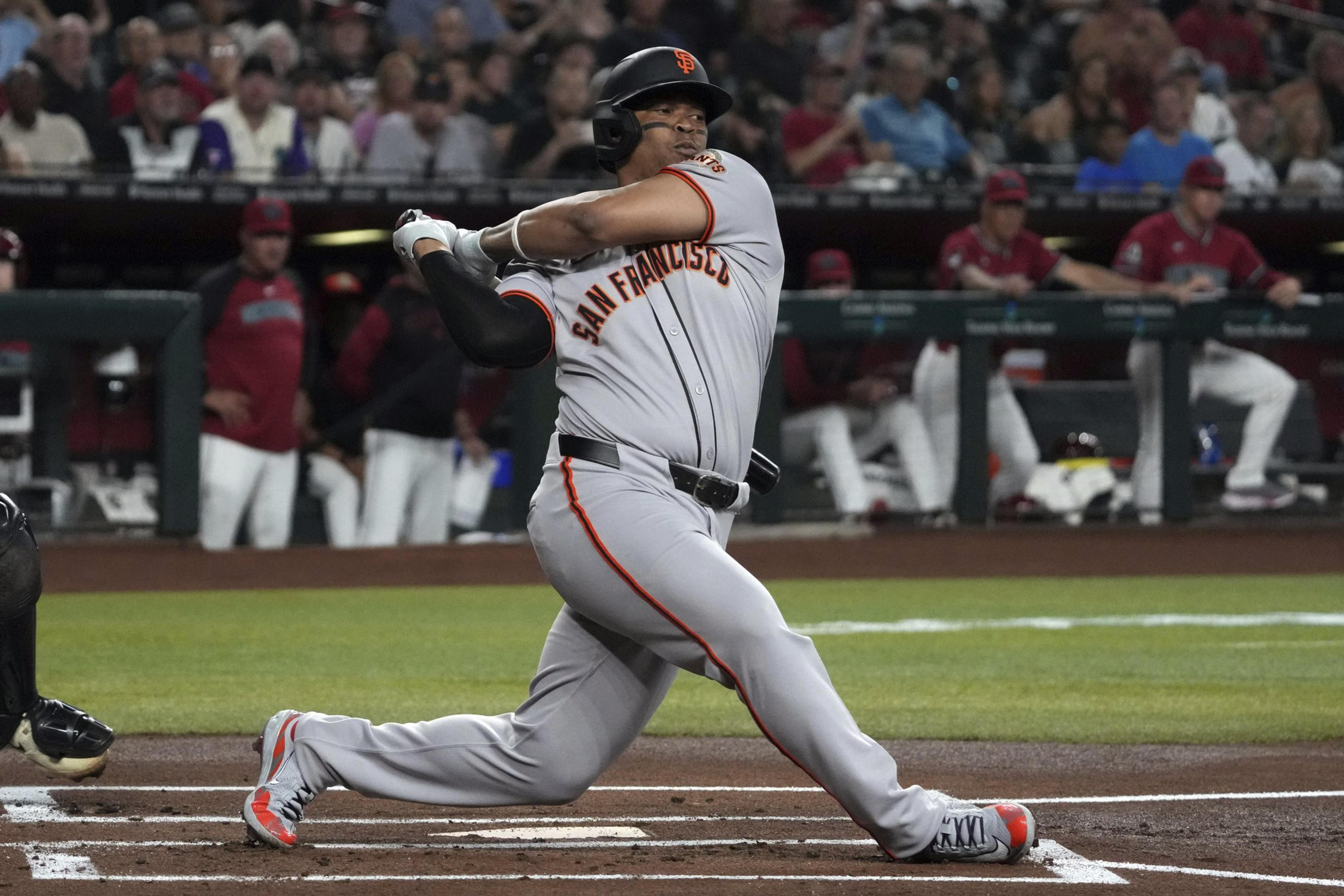 A baseball player in a gray uniform with "San Francisco" swings a bat during a game. The crowd watches from the stands, and teammates sit in the dugout.