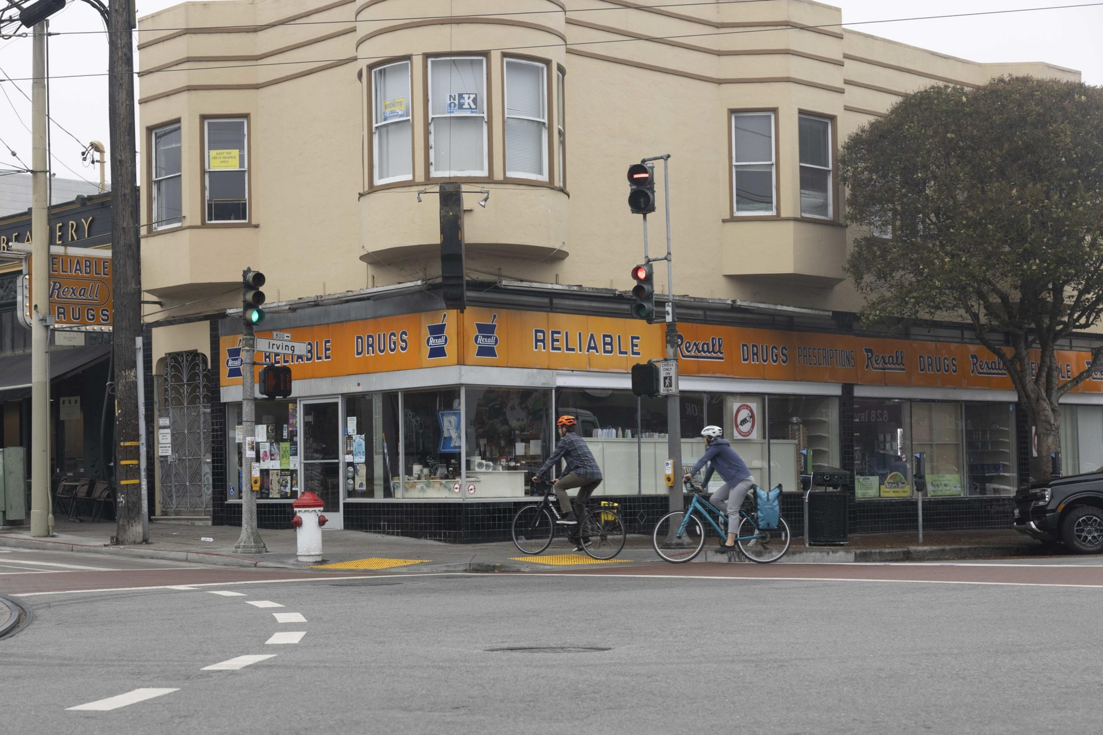 Two cyclists ride past a corner drugstore with an orange and blue &quot;Reliable Drugs&quot; sign. The building is beige with large windows above the storefront.