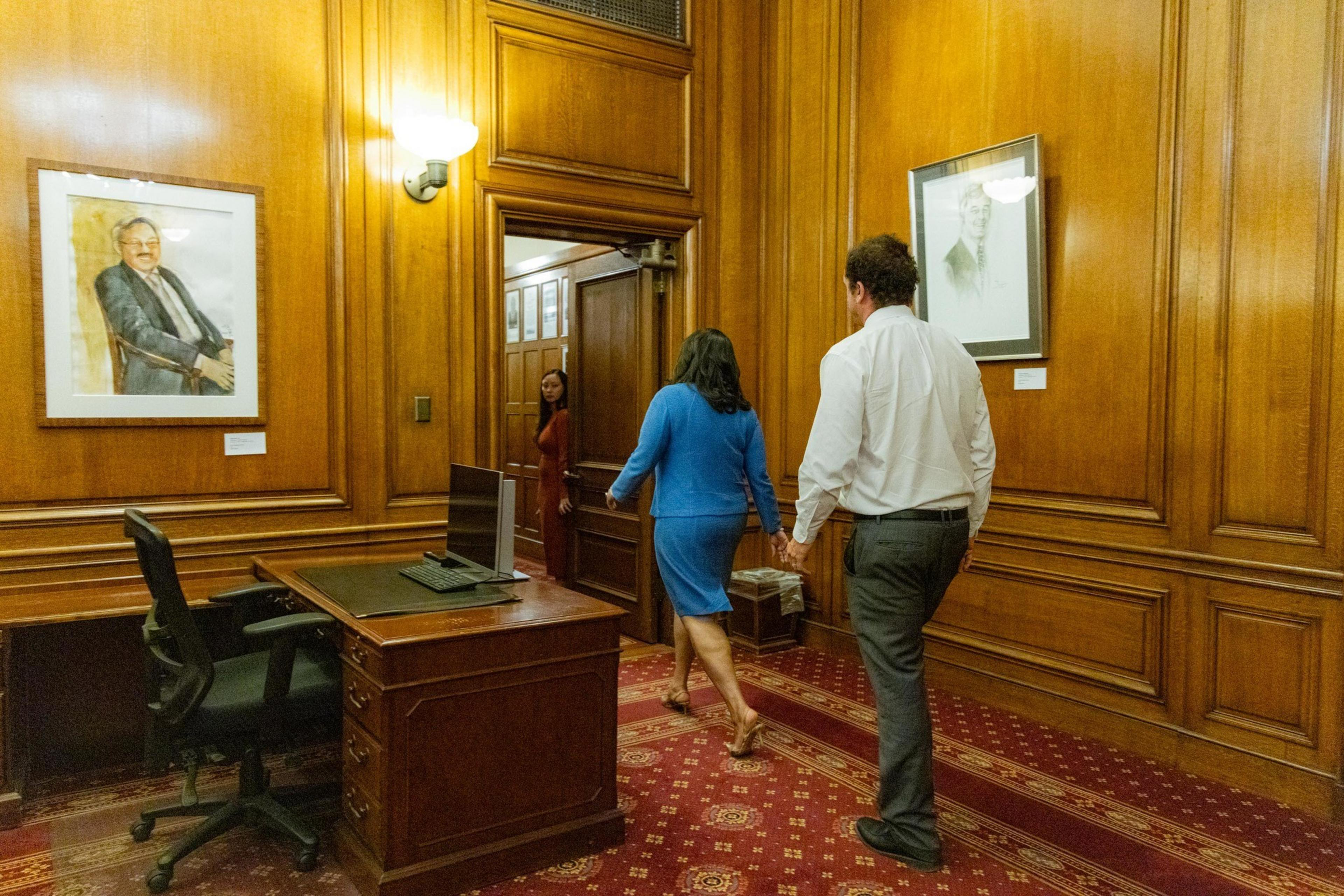 Three people walk through a wood-paneled office with a desk and chairs, adorned with framed portraits on the walls and a red carpet on the floor.
