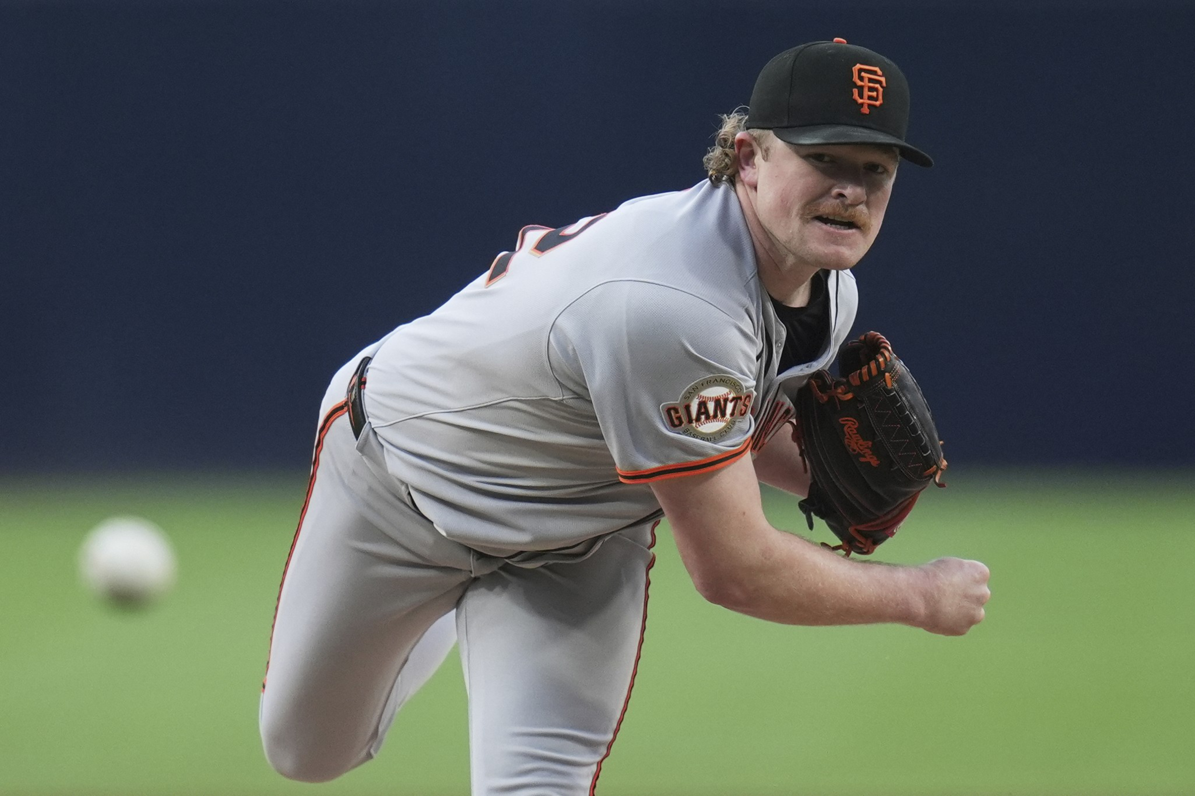 A baseball player in a gray uniform pitches a ball. He wears a black cap with an orange logo and a matching glove, with a focused expression on his face.