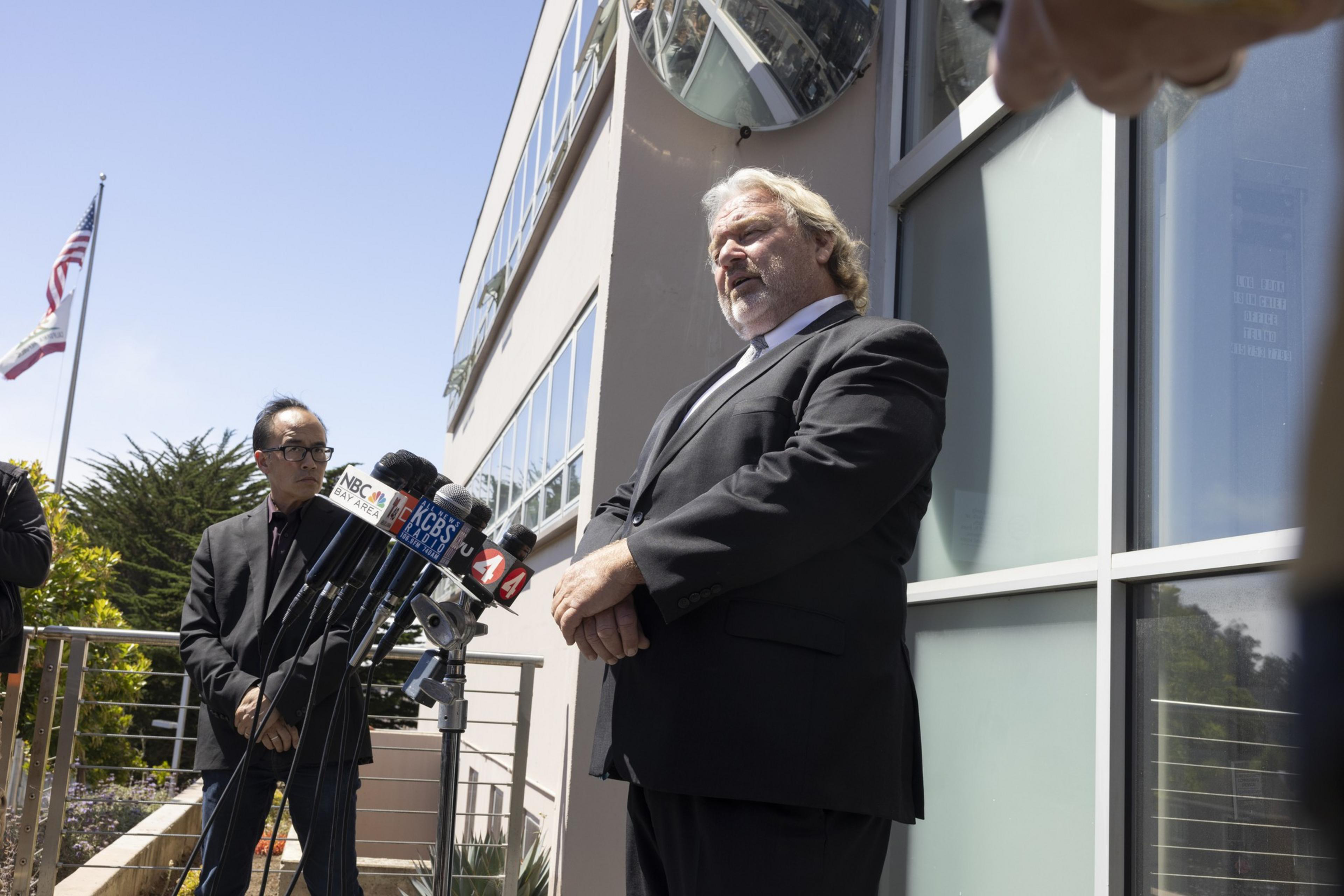 A man in a black suit speaks at a podium with multiple microphones outside a building. Another person stands beside him, and flags are in the background.