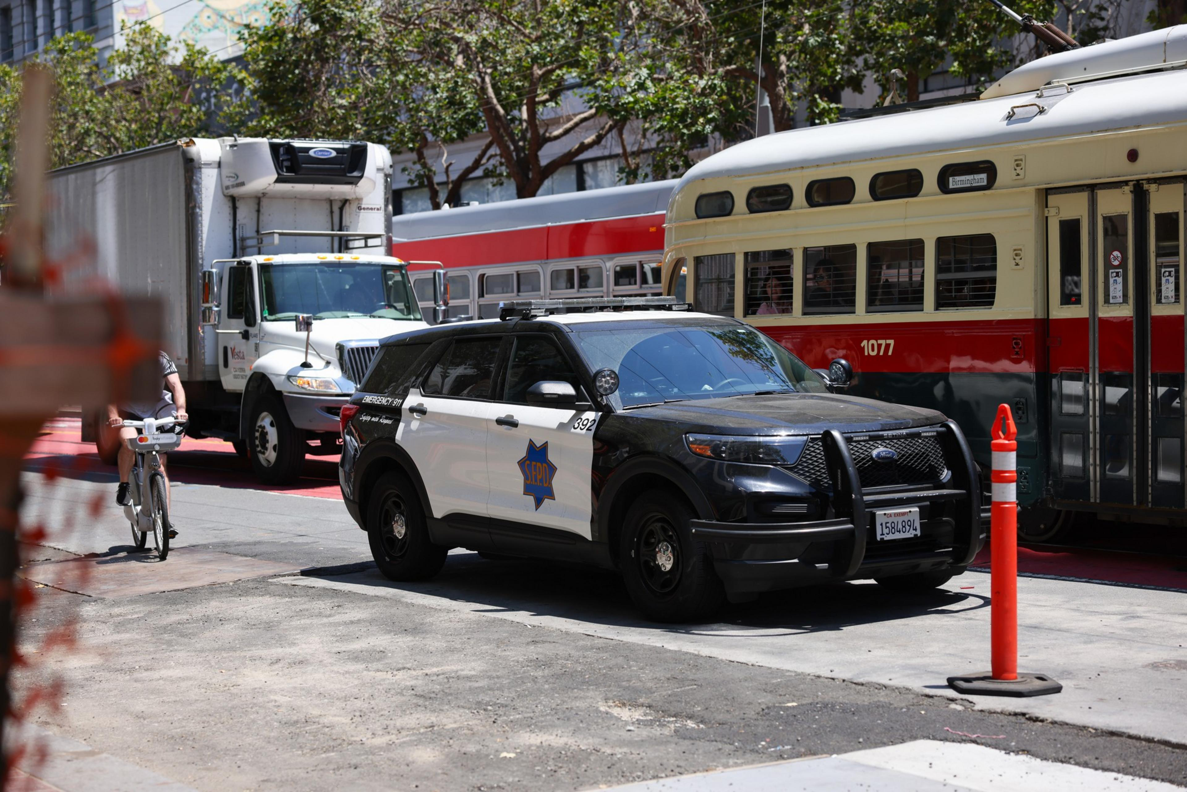 A black and white police SUV is parked on the street next to a red and cream tram, a white delivery truck, and a cyclist passing by.