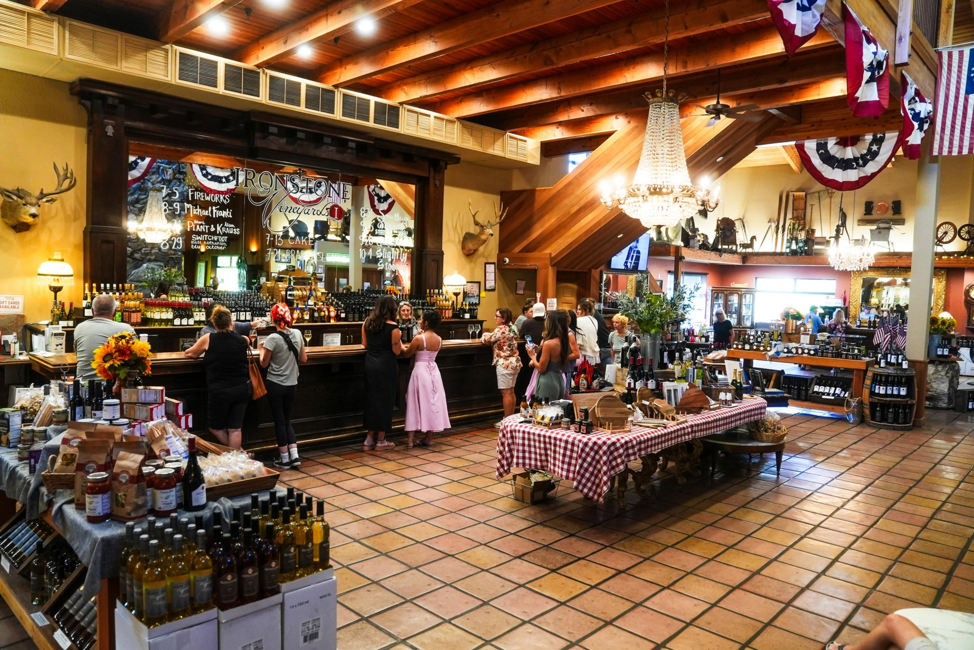 The image shows a cozy wine tasting room with a wooden bar, patrons enjoying wine, and tables displaying bottles and packaged goods under warm lighting.