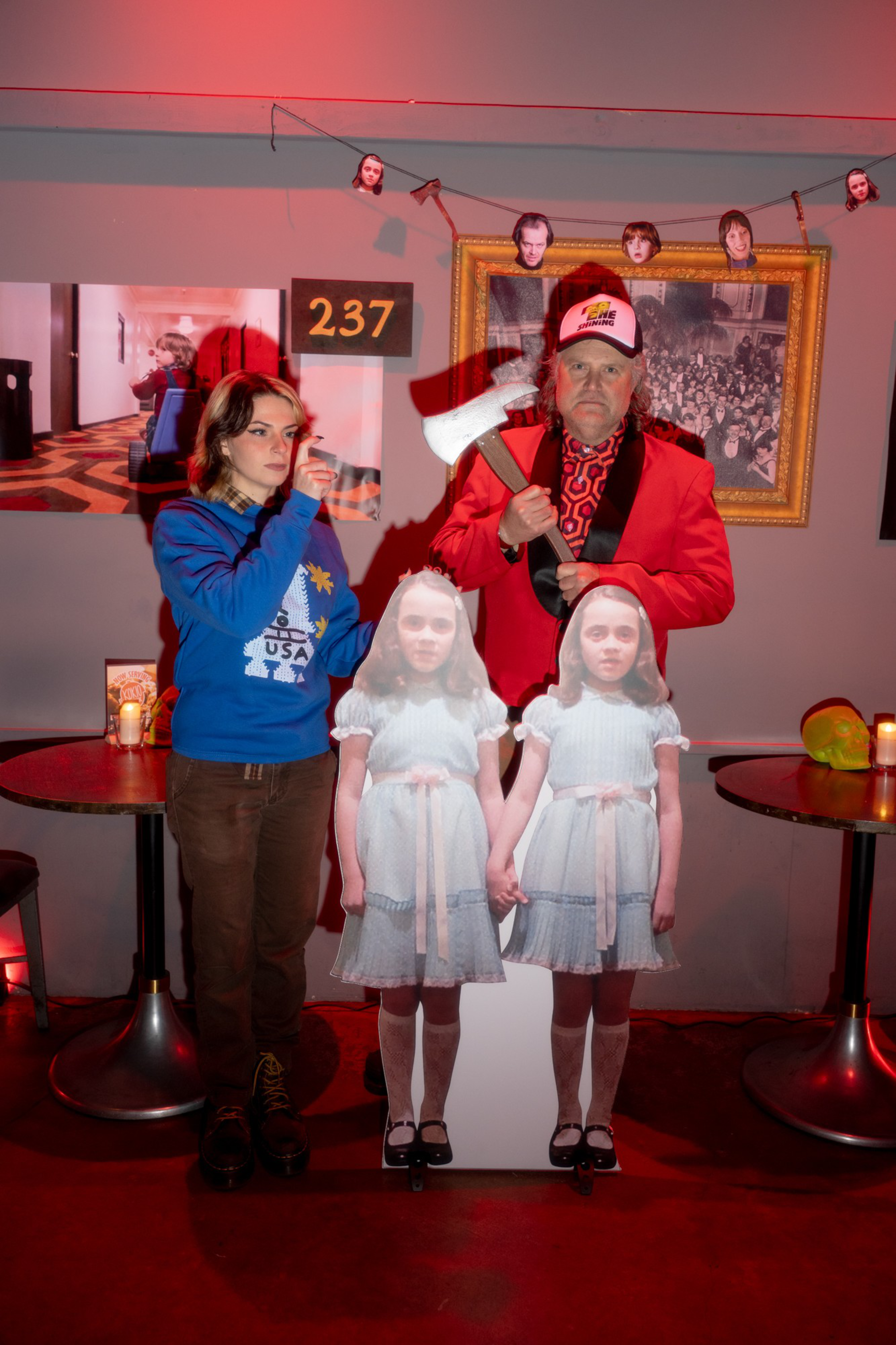 A woman points a finger gun as a man in a red jacket holds an axe, standing behind a cutout of the twin girls from "The Shining" in a dimly lit room.