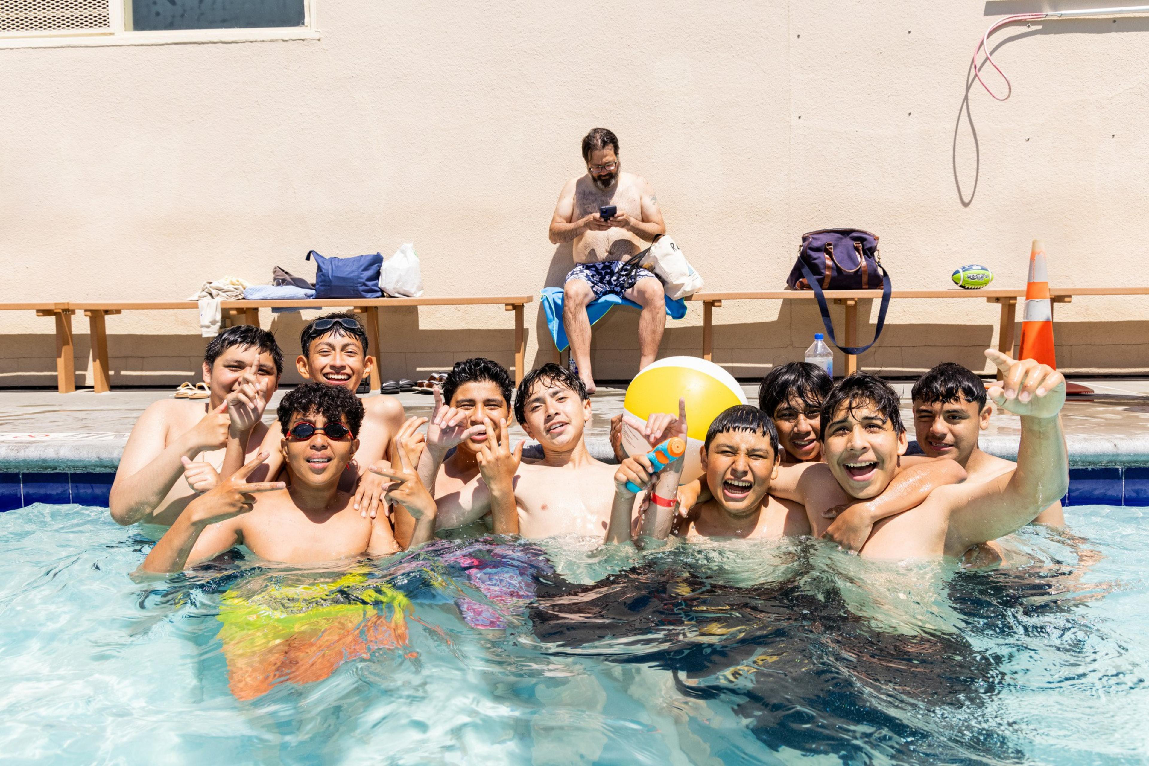 A group of boys is joyfully posing in a swimming pool, some holding a ball and a water gun, with a man sitting on a bench in the background using his phone.