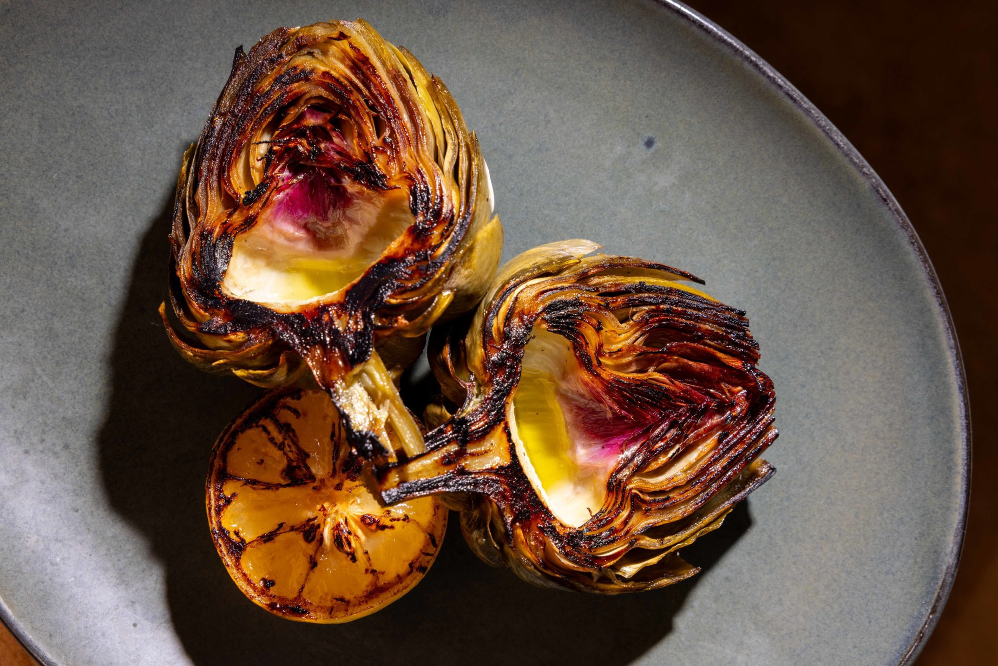 Two charred artichoke halves and a grilled lemon half rest on a gray plate against a dark background.
