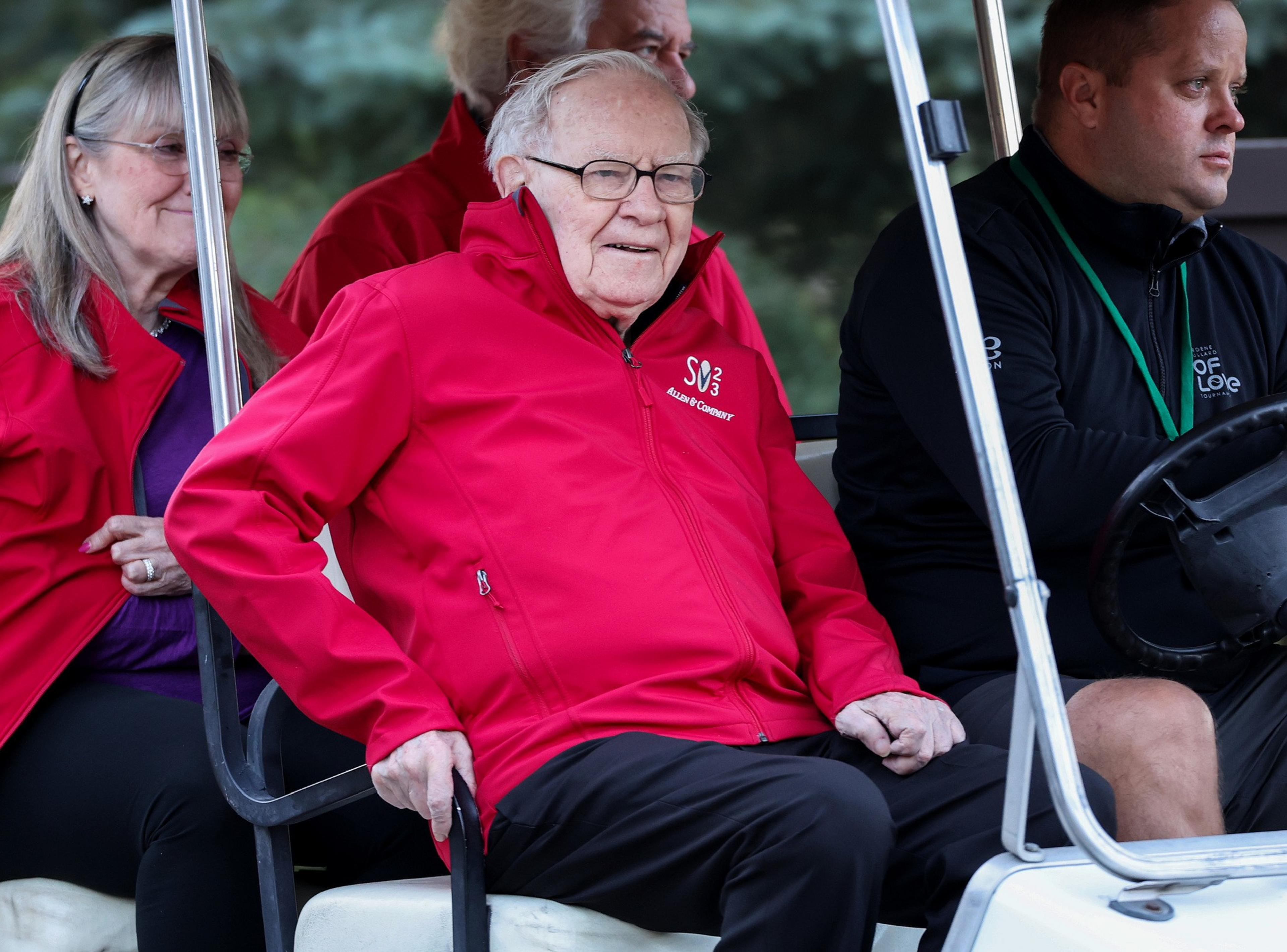 An elderly man in a red jacket sits on a golf cart, accompanied by a woman and a driver. The background is blurred with a hint of greenery.
