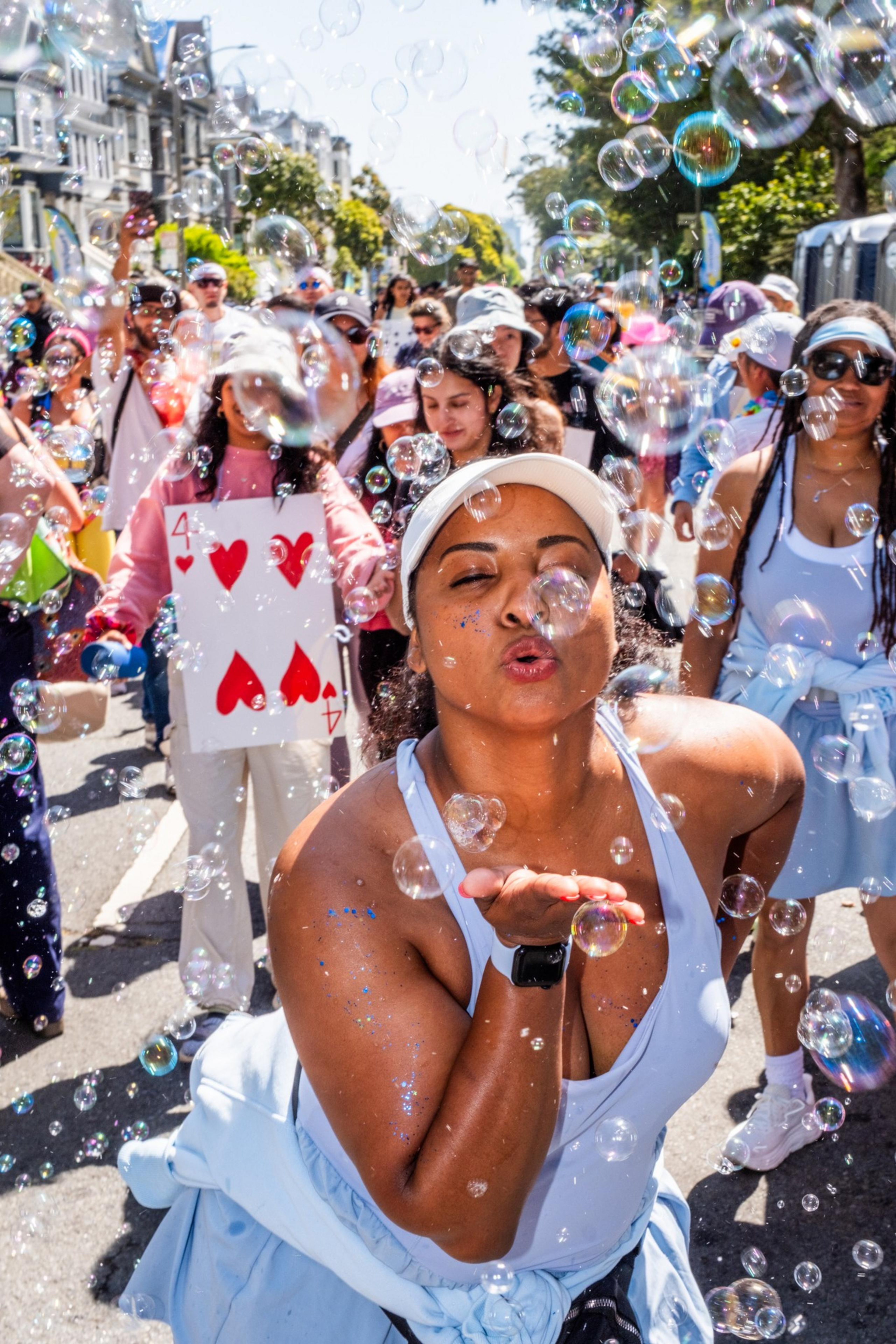 A joyful woman wearing a cap blows bubbles surrounded by a lively crowd. People in playful outfits, including a person holding a large card, enjoy the sunny street.