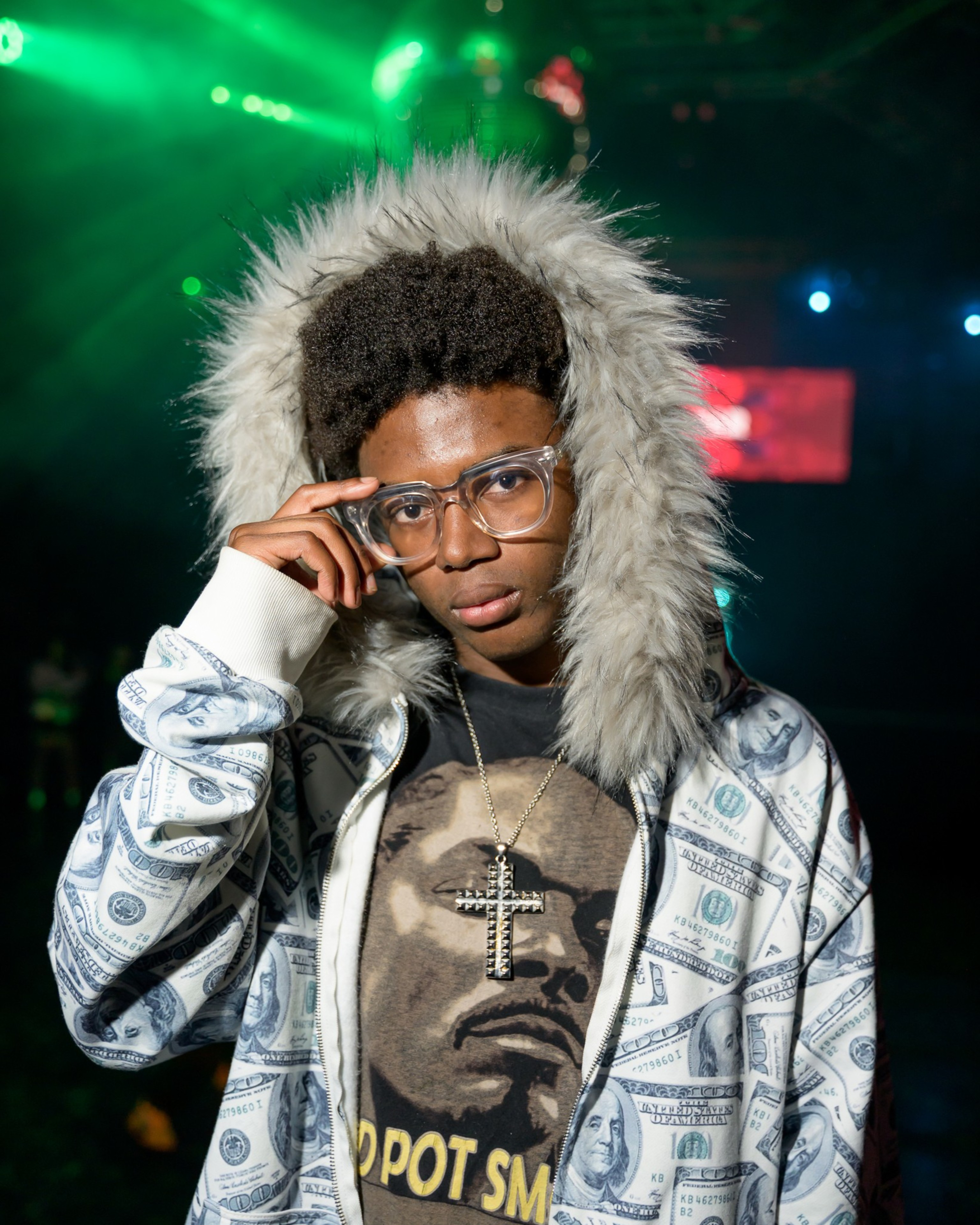 A person in glasses wears a fur-hooded jacket printed with dollar bills and a graphic T-shirt. The background features colorful stage lights.