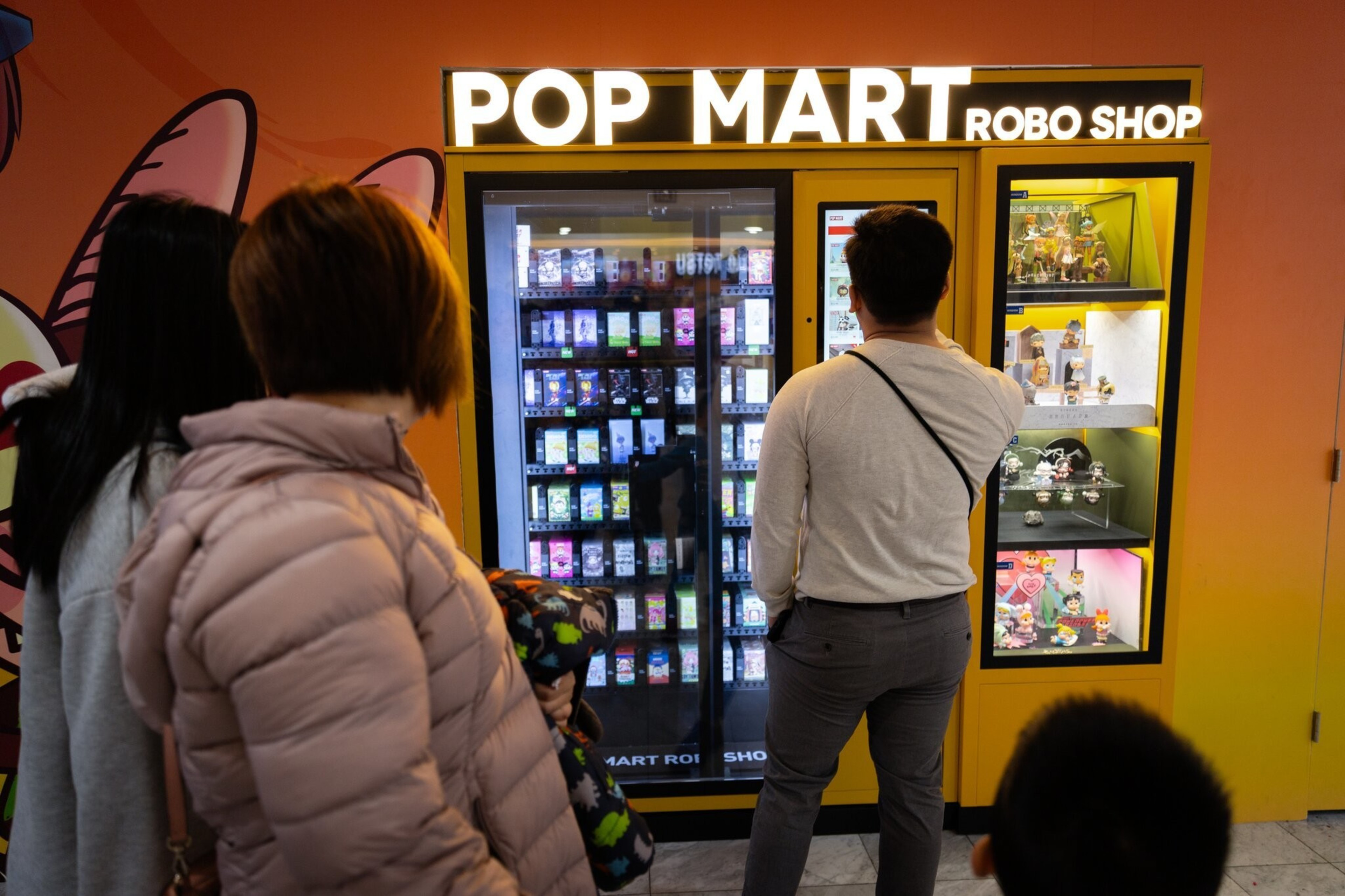 A group of people stand in front of a brightly lit vending machine labeled &quot;POP MART ROBO SHOP,&quot; which dispenses various small collectible items.