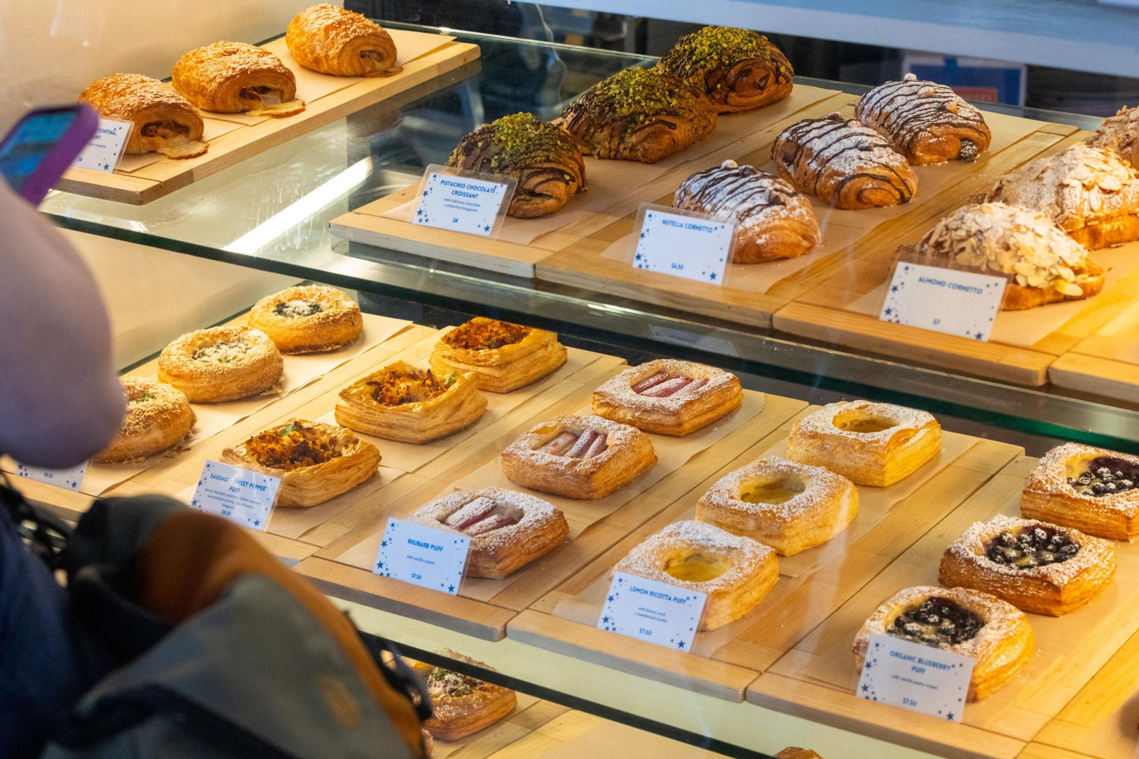 A bakery display case holds various pastries including croissants with nuts, chocolate, and powdered sugar, and square Danish pastries topped with fruit and nuts.