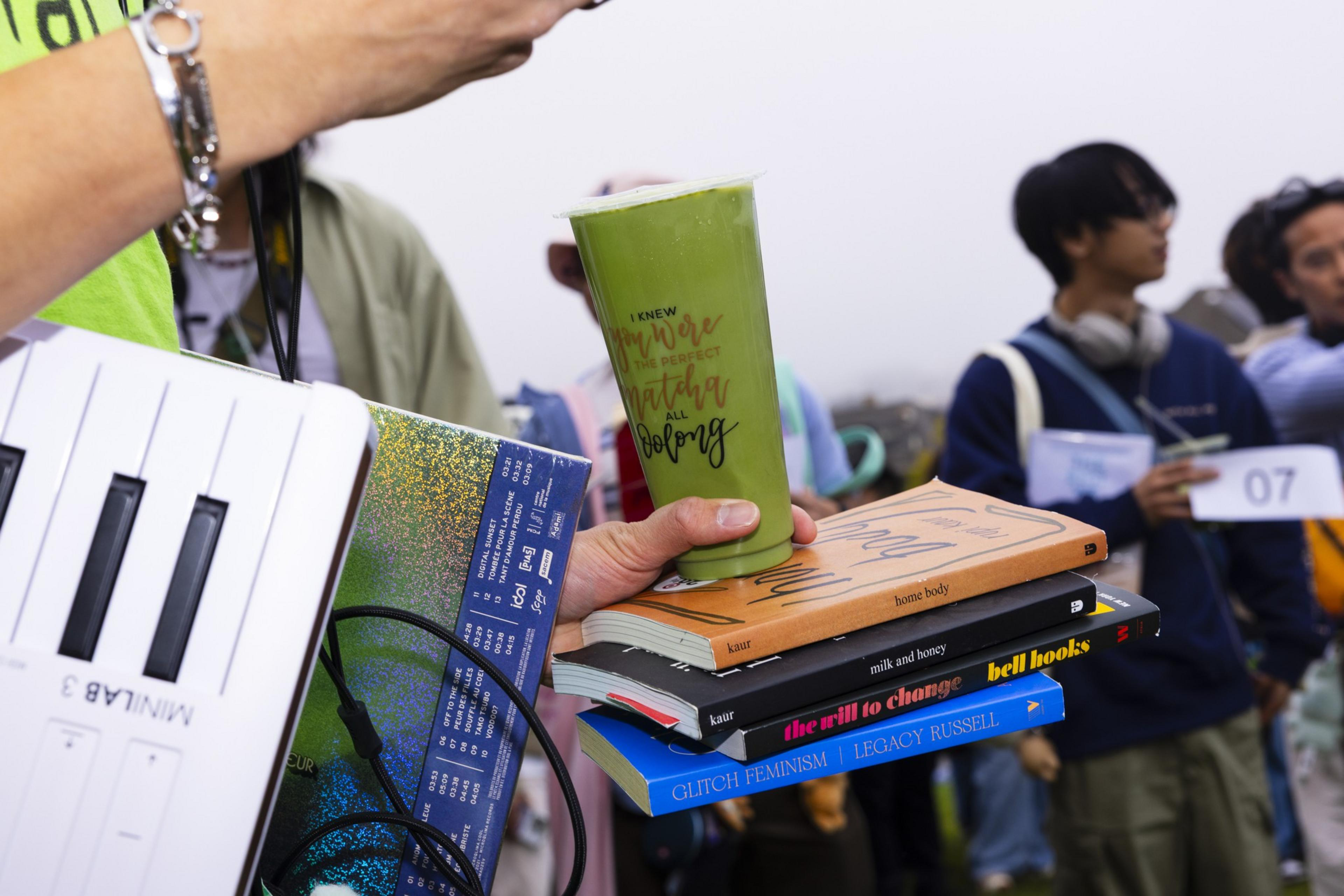 A person holds a green drink and a stack of four books while carrying a white MIDI keyboard. Other people stand in the blurred background.