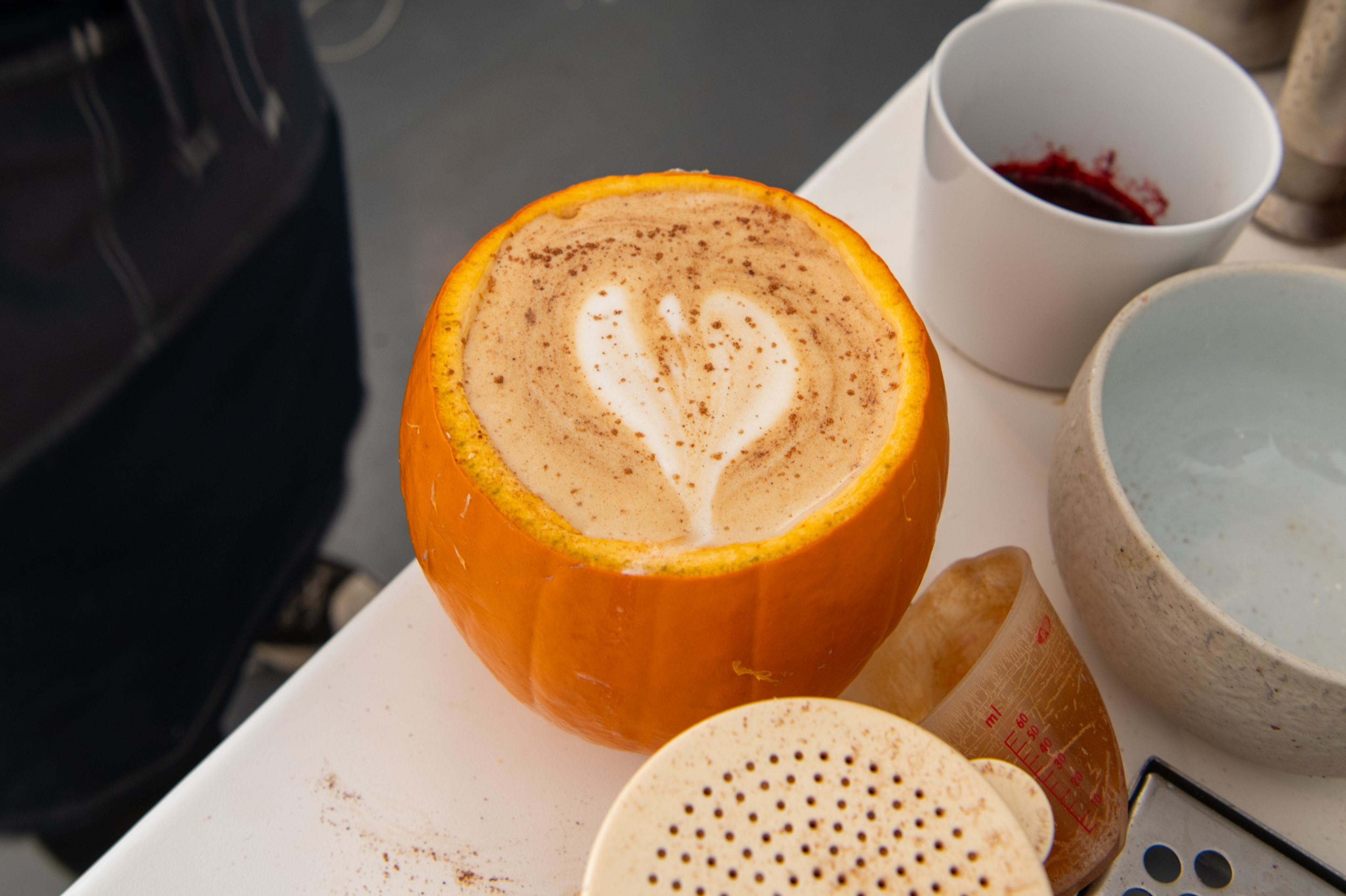 A pumpkin hollowed out and used as a cup holds a latte with heart-shaped foam art on top, surrounded by kitchen items on a white surface.