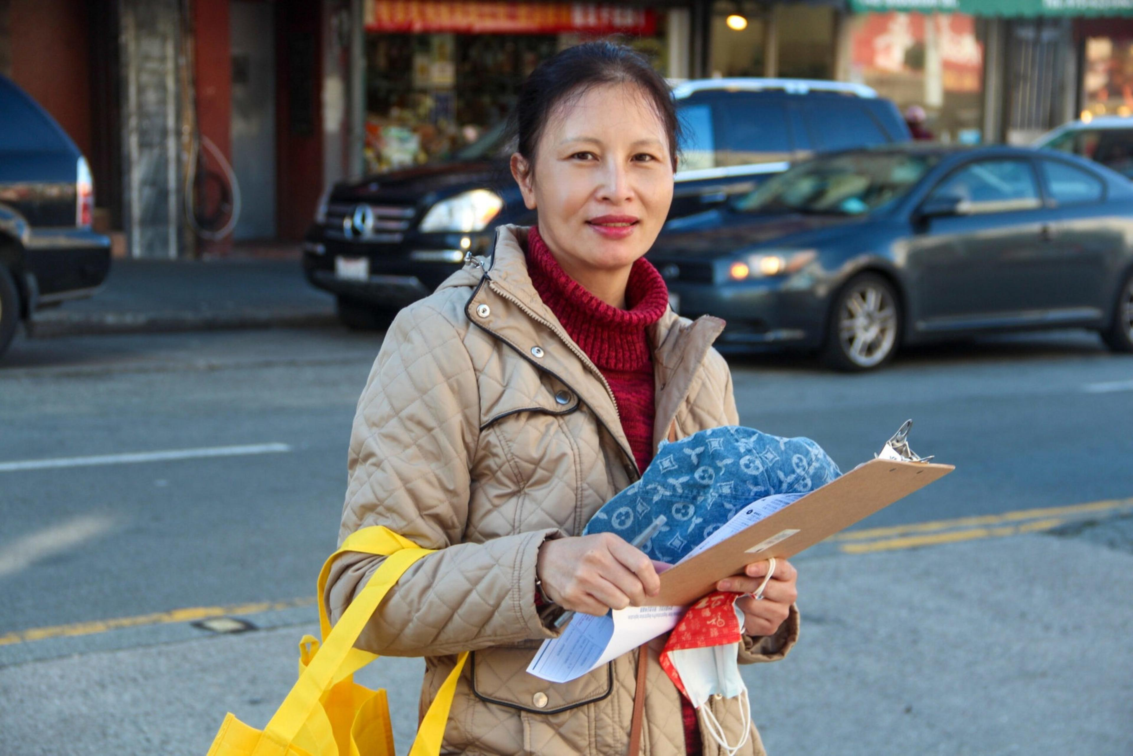 A woman in a beige quilted jacket and red sweater holds a clipboard, papers, and yellow reusable bags while standing on a street with cars behind her.