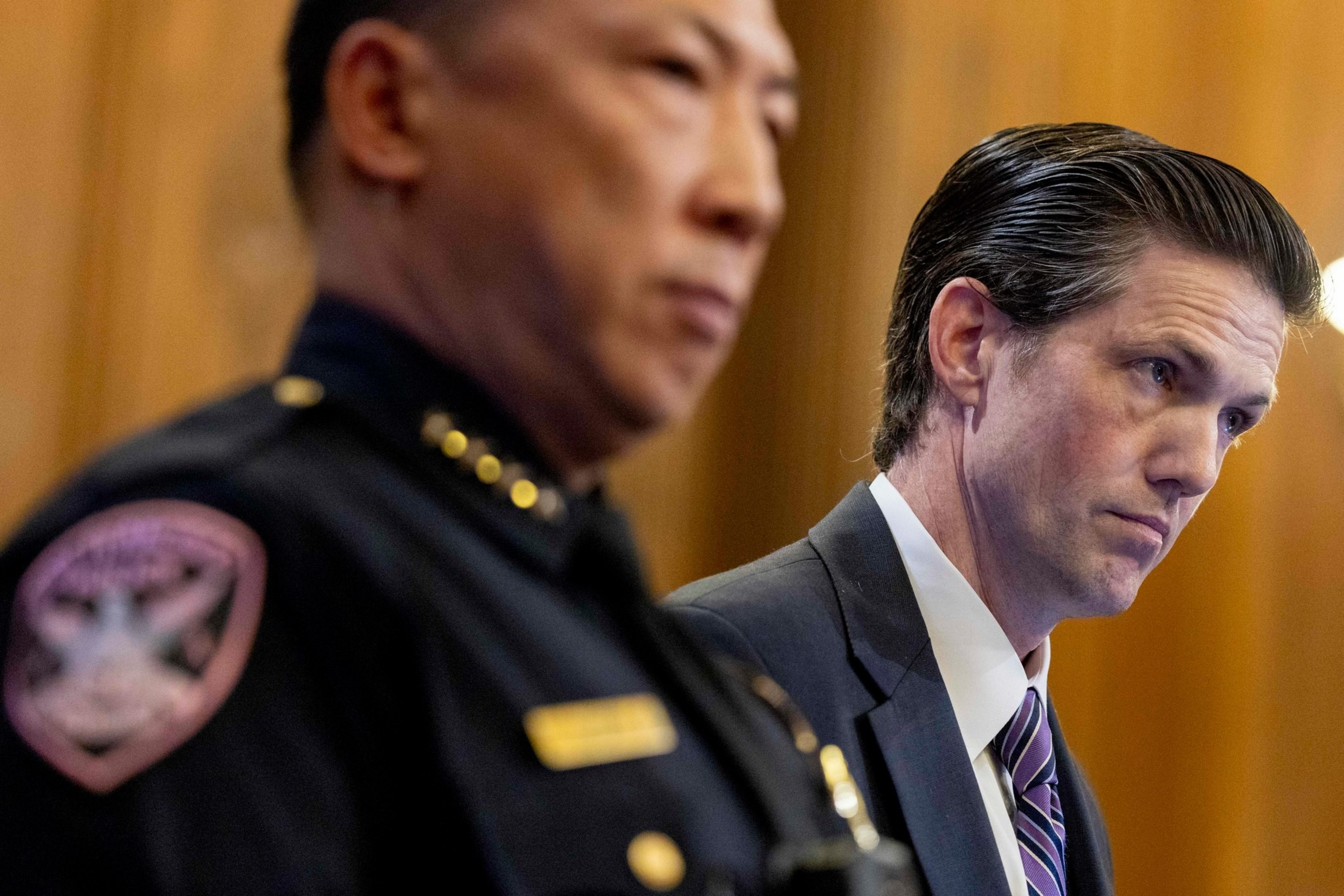 A police officer in uniform and a man in a suit with a striped tie stand side by side, both looking serious and focused.