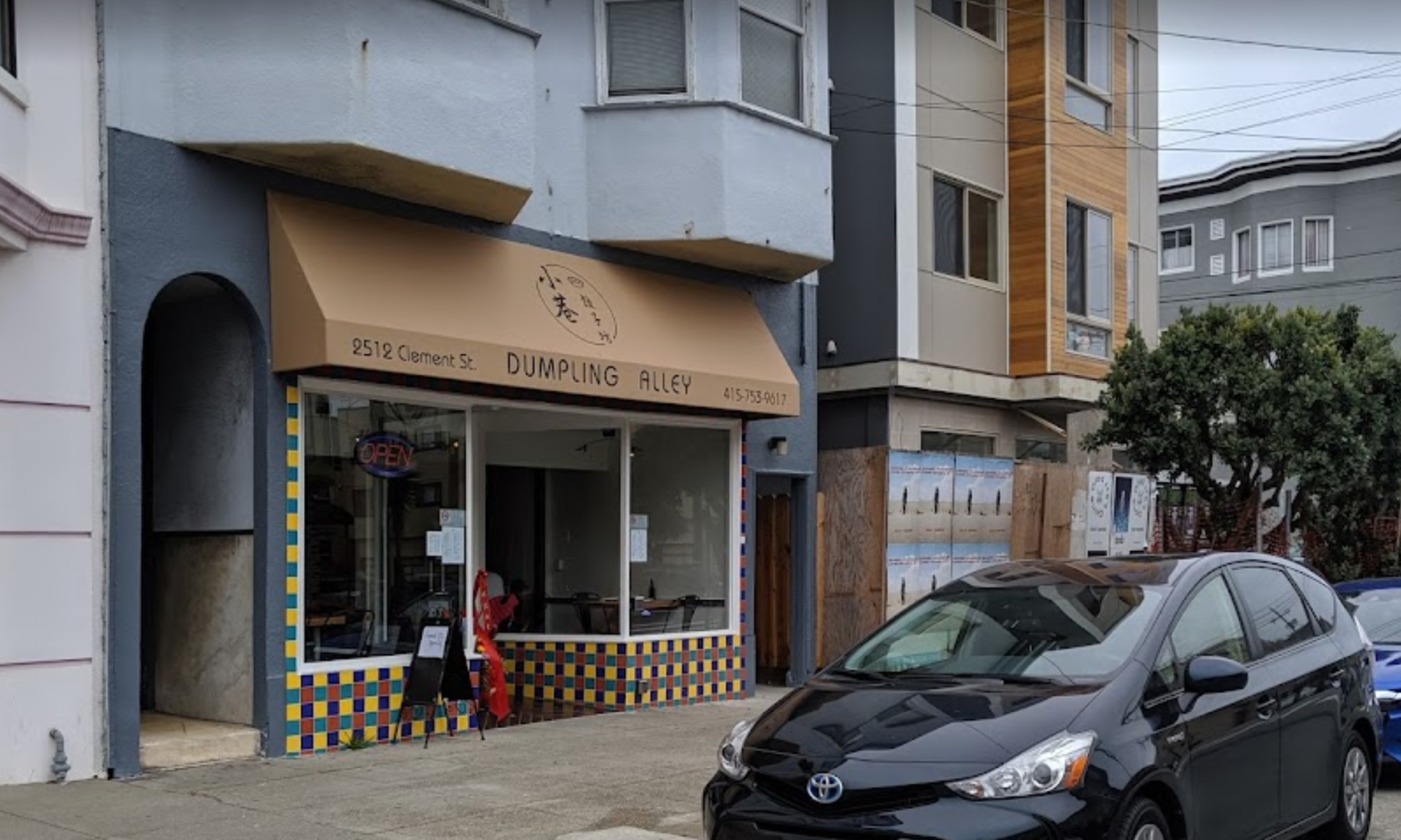 A small restaurant named Dumpling Alley with a beige awning and colorful tile base is next to a sidewalk where a black Toyota car is parked.