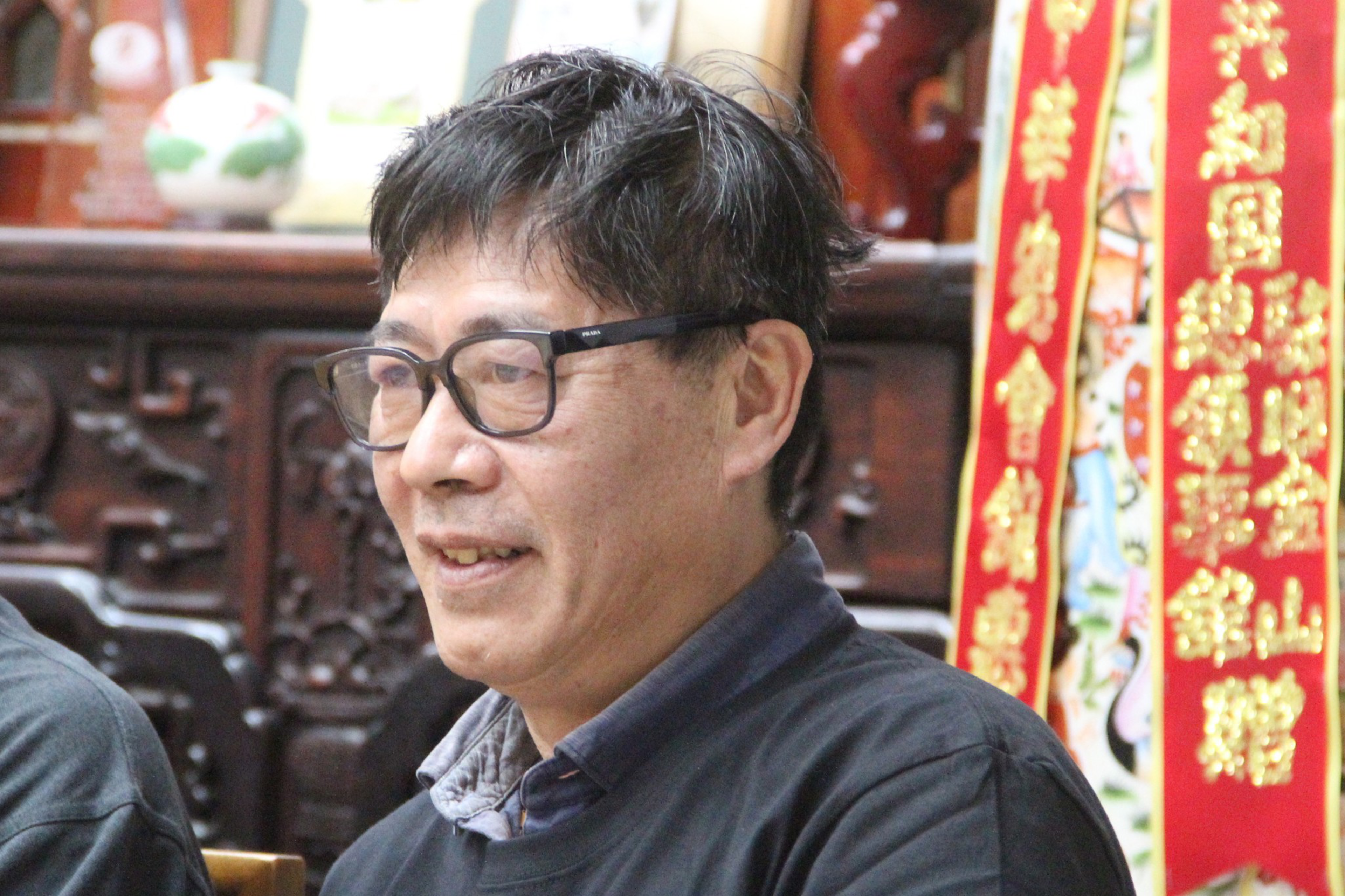 A middle-aged man with glasses wearing a dark shirt is smiling slightly, with red and gold Chinese decorations in the background.
