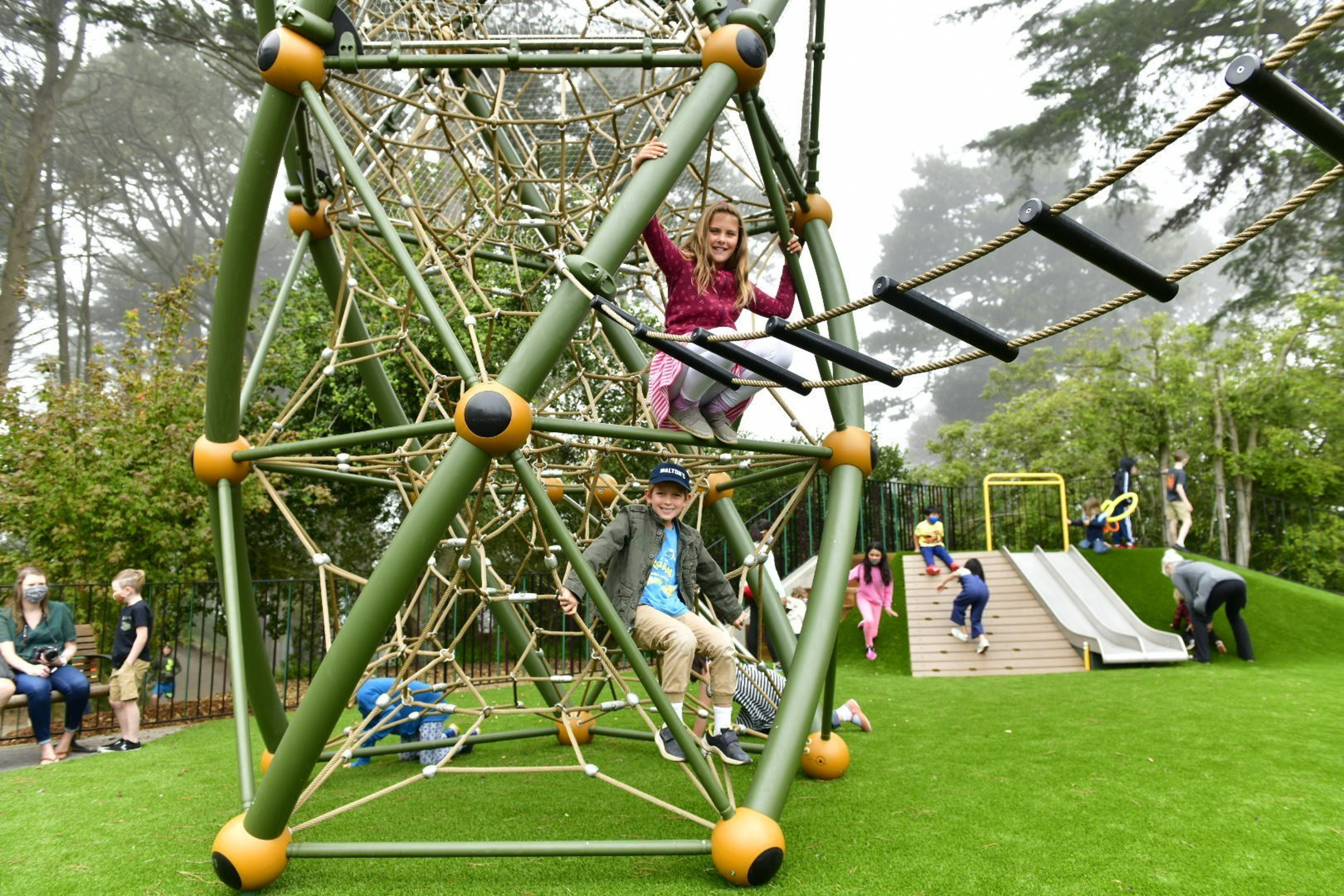 Two children climb on a large green and beige rope pyramid structure in a playground with slides and other kids playing nearby.