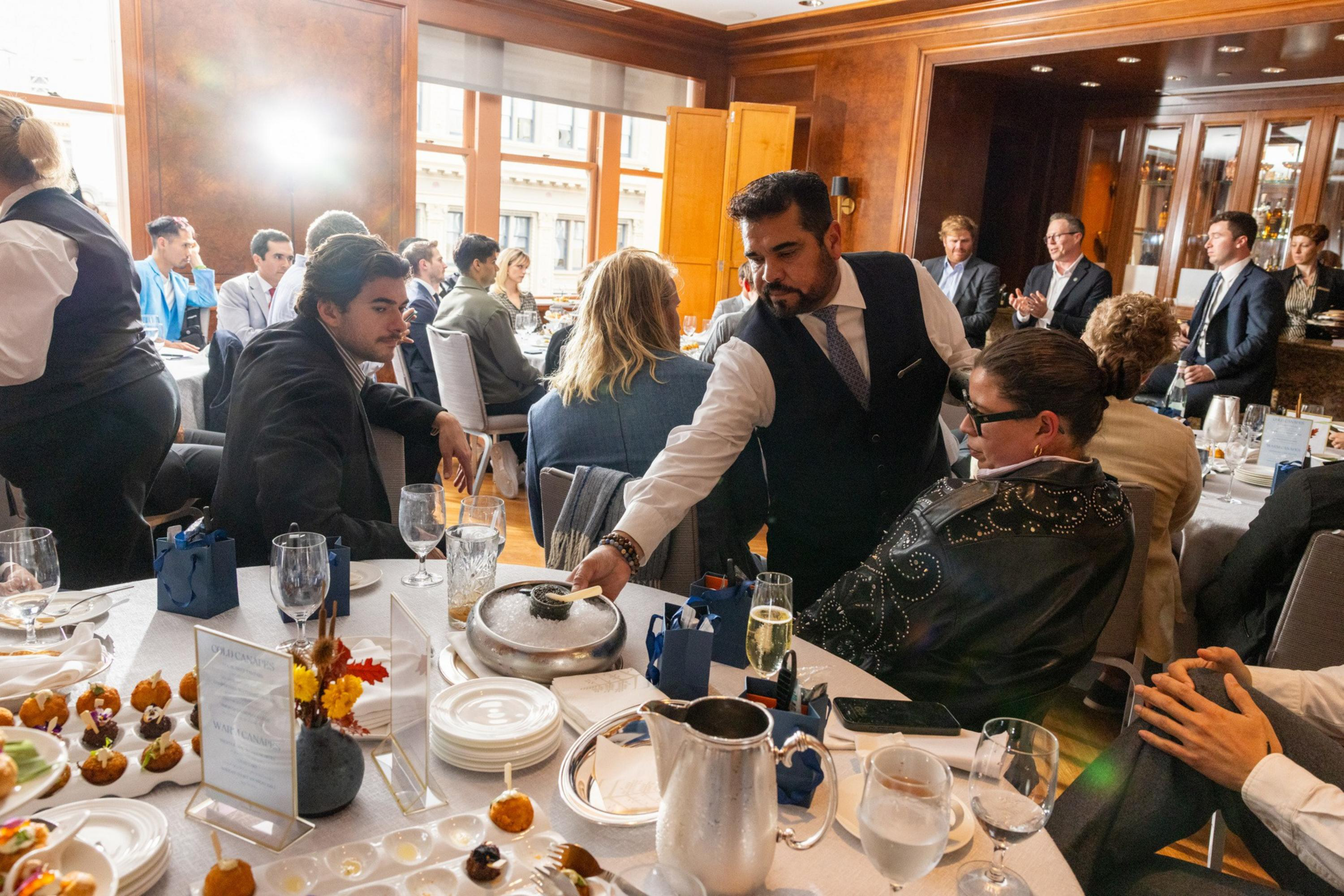 A waiter serves food to guests seated around a table set with drinks and appetizers in a formal dining room.