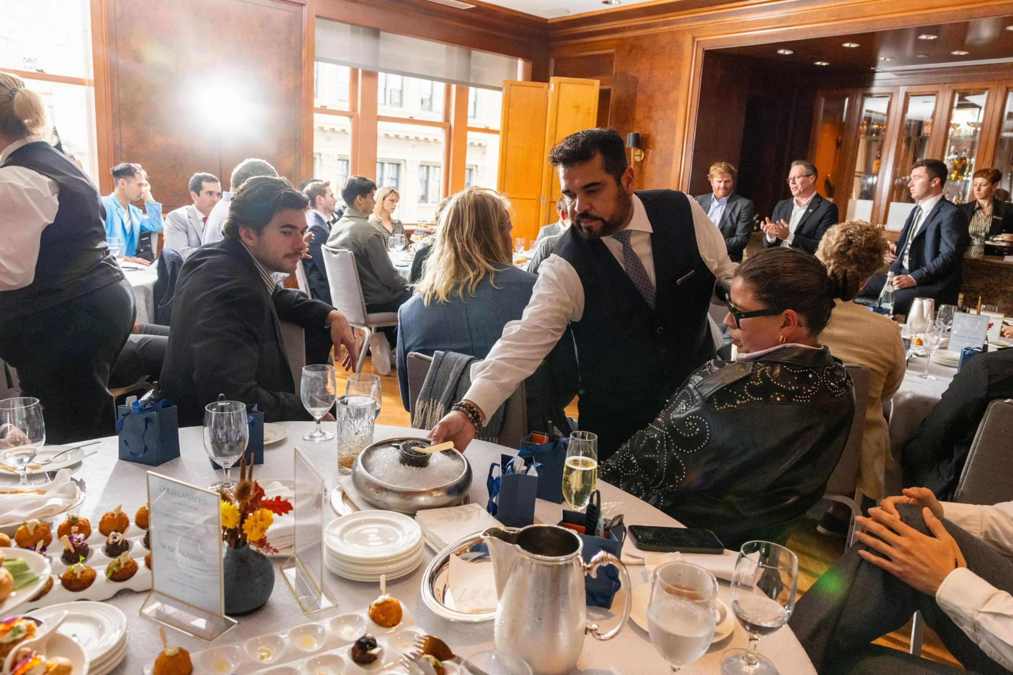 A waiter serves food to guests seated around a table set with drinks and appetizers in a formal dining room.