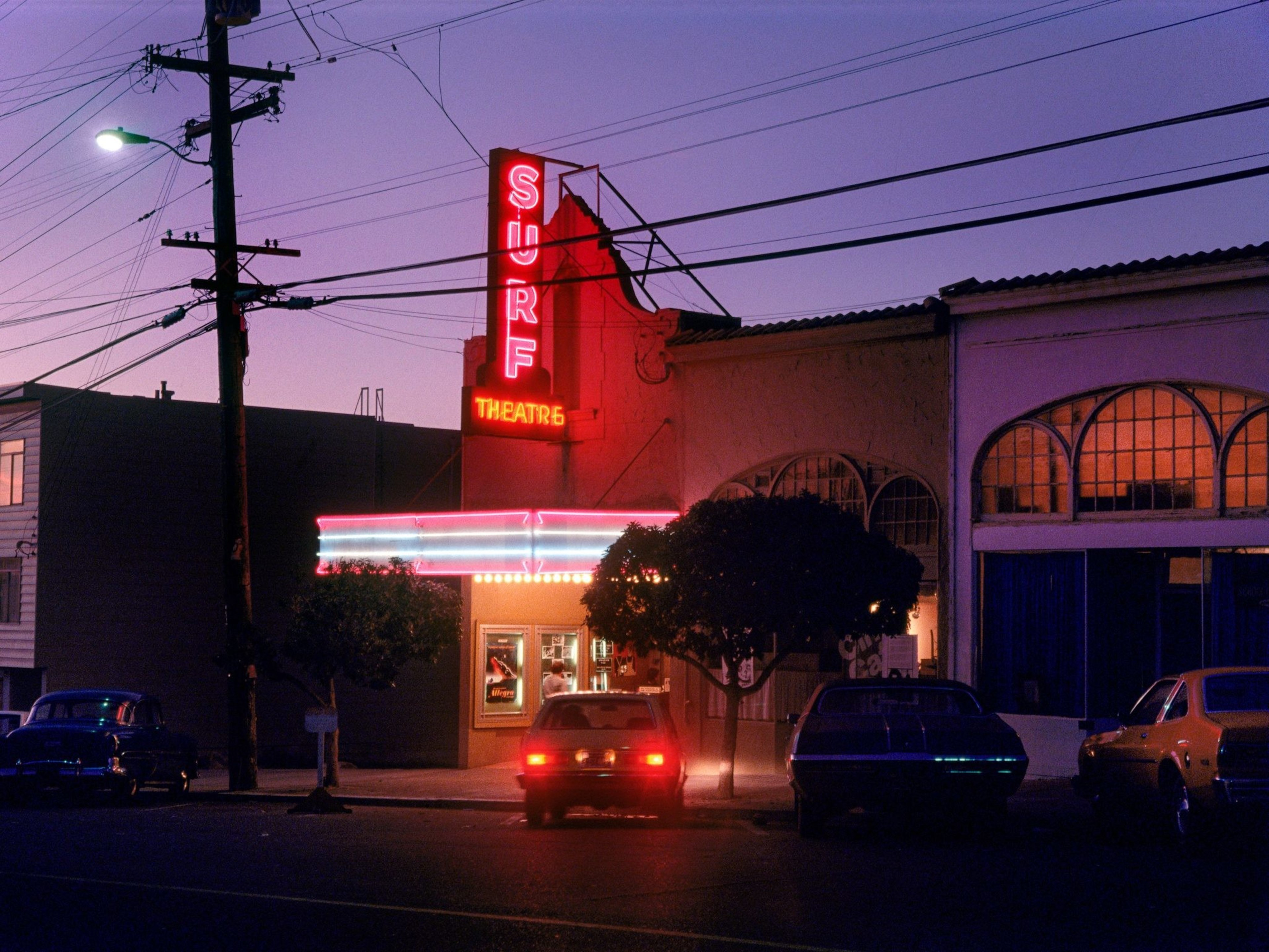 The image shows a vintage theater at dusk with a neon sign reading &quot;Surf Theatre.&quot; Several cars are parked in front, and trees are outlined against a deep purple sky.