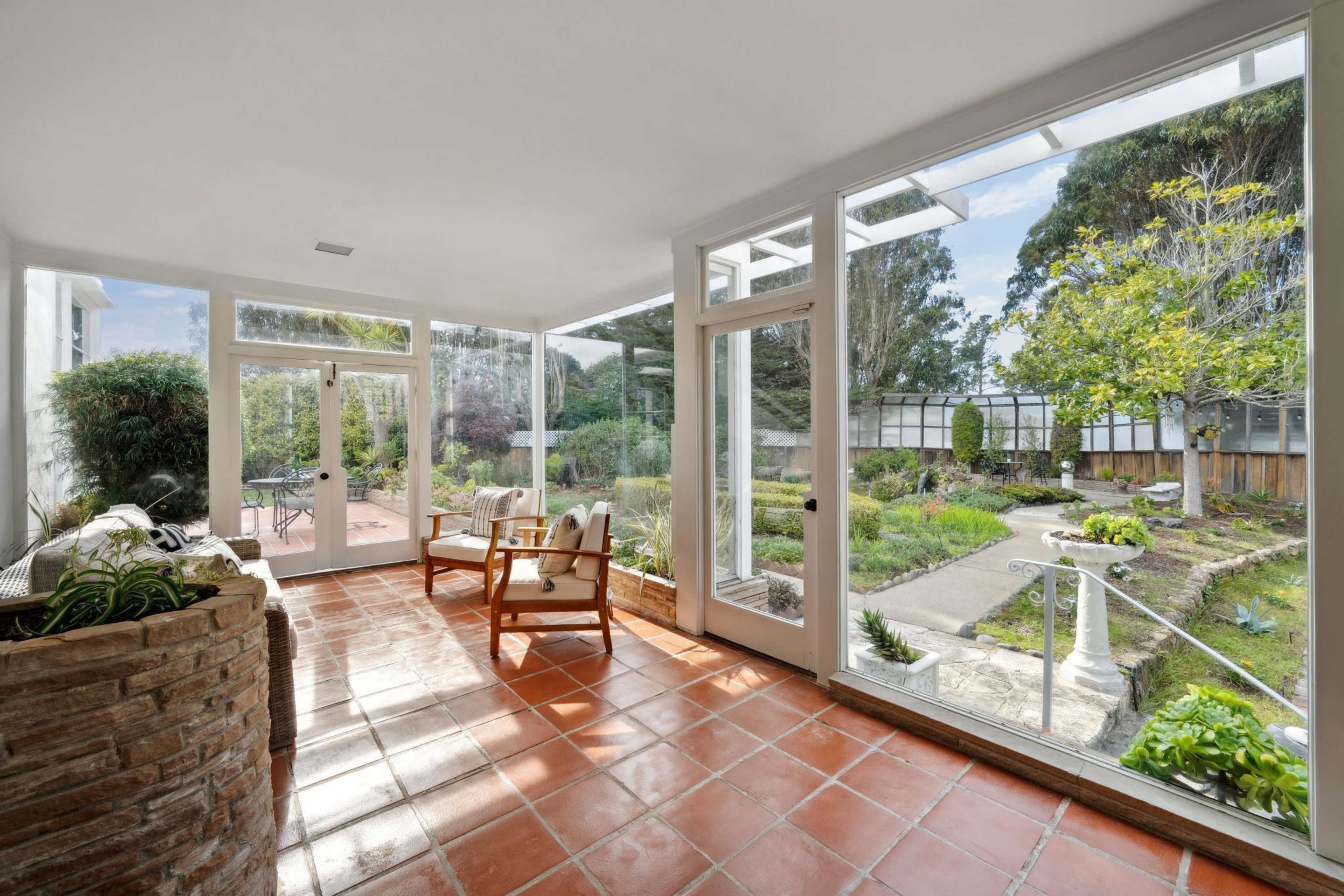 A sunlit room with terracotta tile flooring and glass walls opens to a garden with a stone pathway, leafy trees, plants, and outdoor seating.