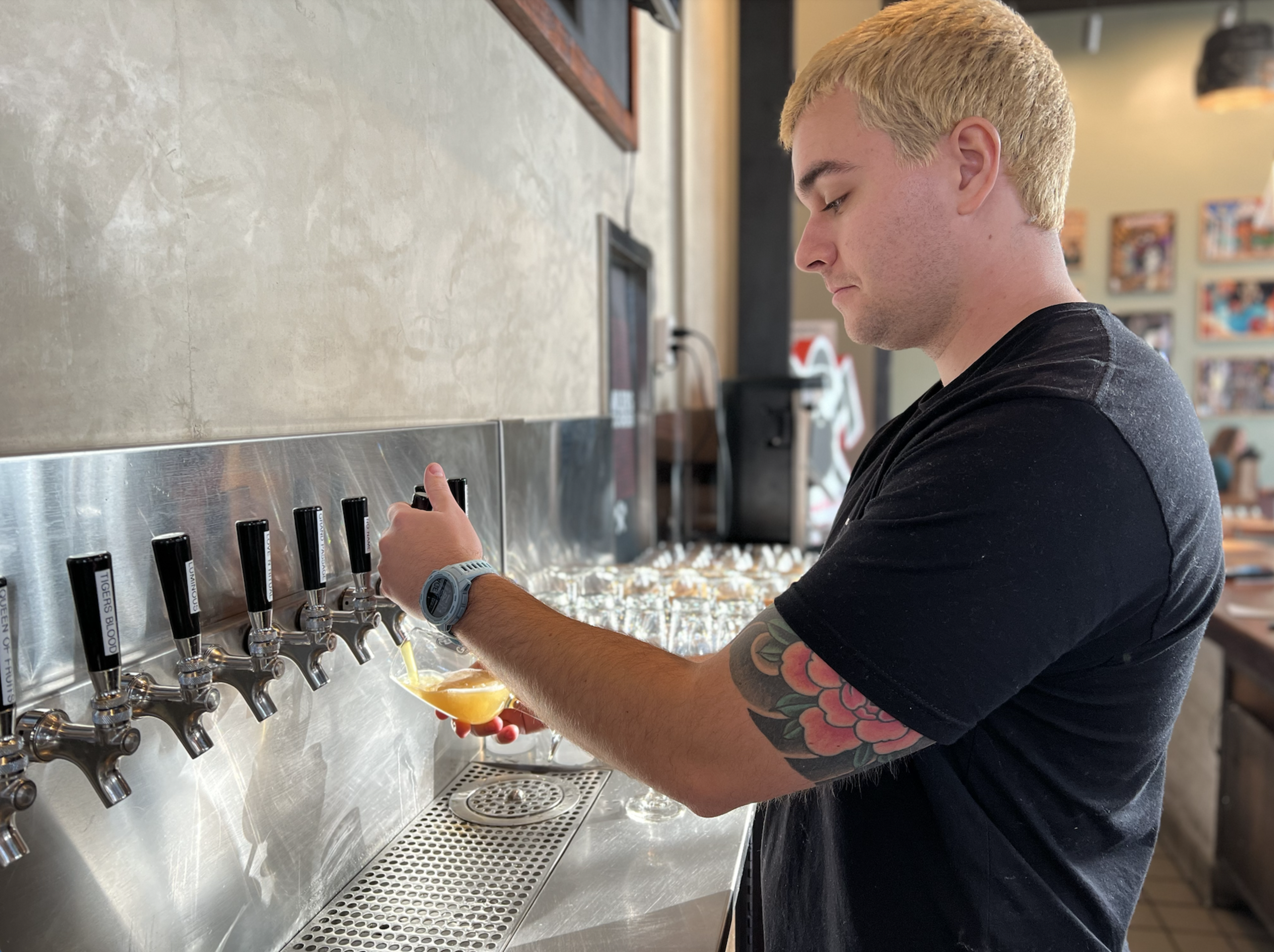 A man with a blonde buzz cut and a floral tattoo on his arm pours a drink from a tap into a glass at a bar with multiple taps.