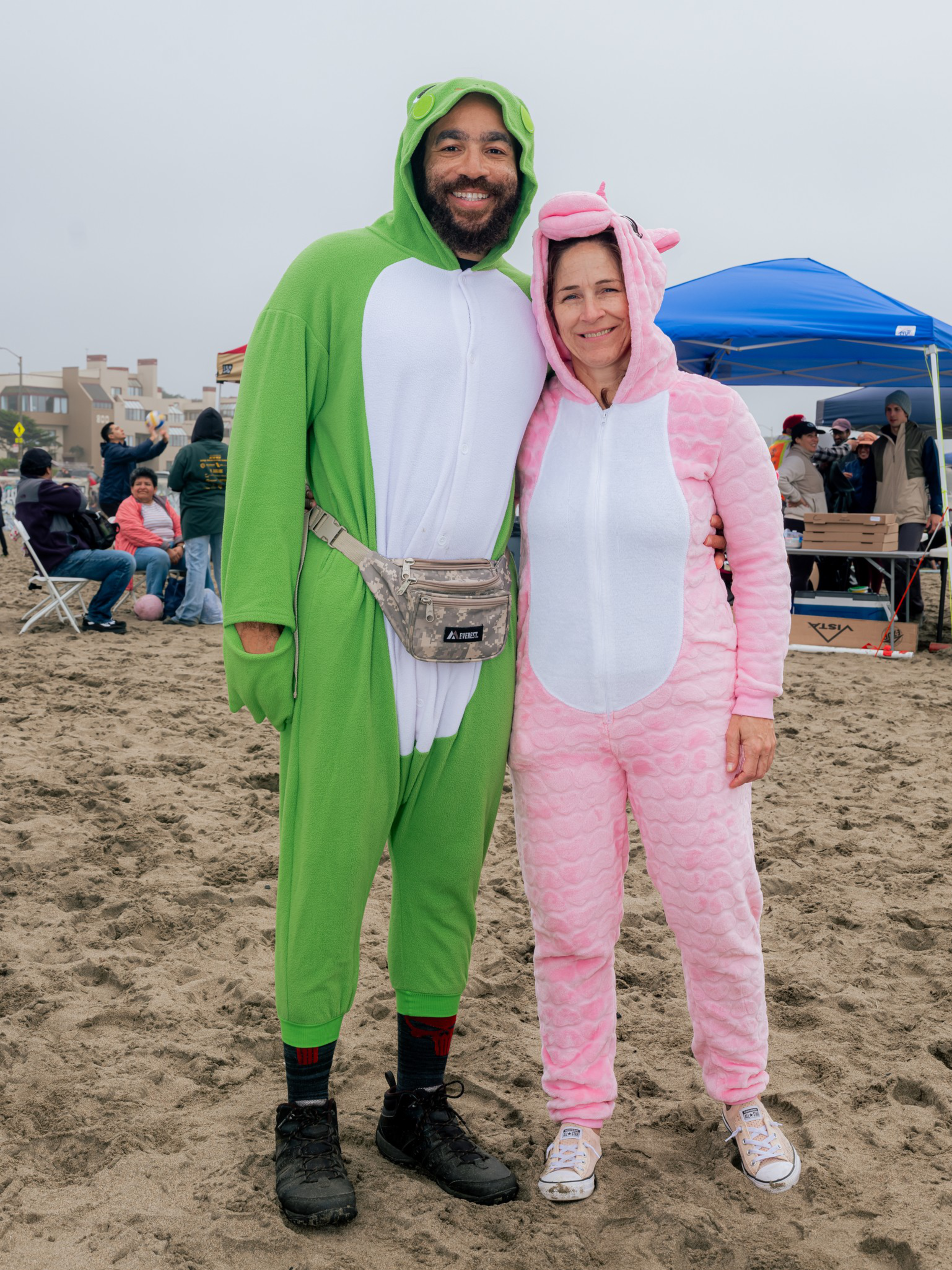 A man and a woman stand on sandy ground wearing full-body animal onesies, the man in green and white and the woman in pink, both smiling warmly.