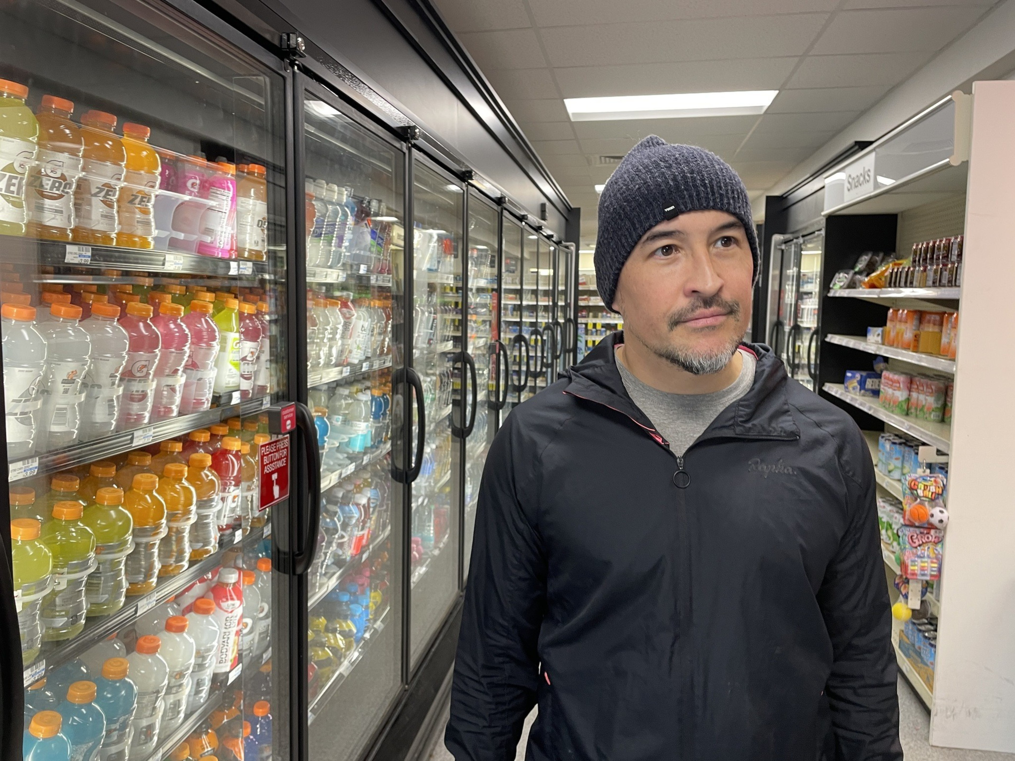 A man in a black jacket and beanie stands in a store aisle with various drinks in refrigerated cases on the left and snacks on the right.