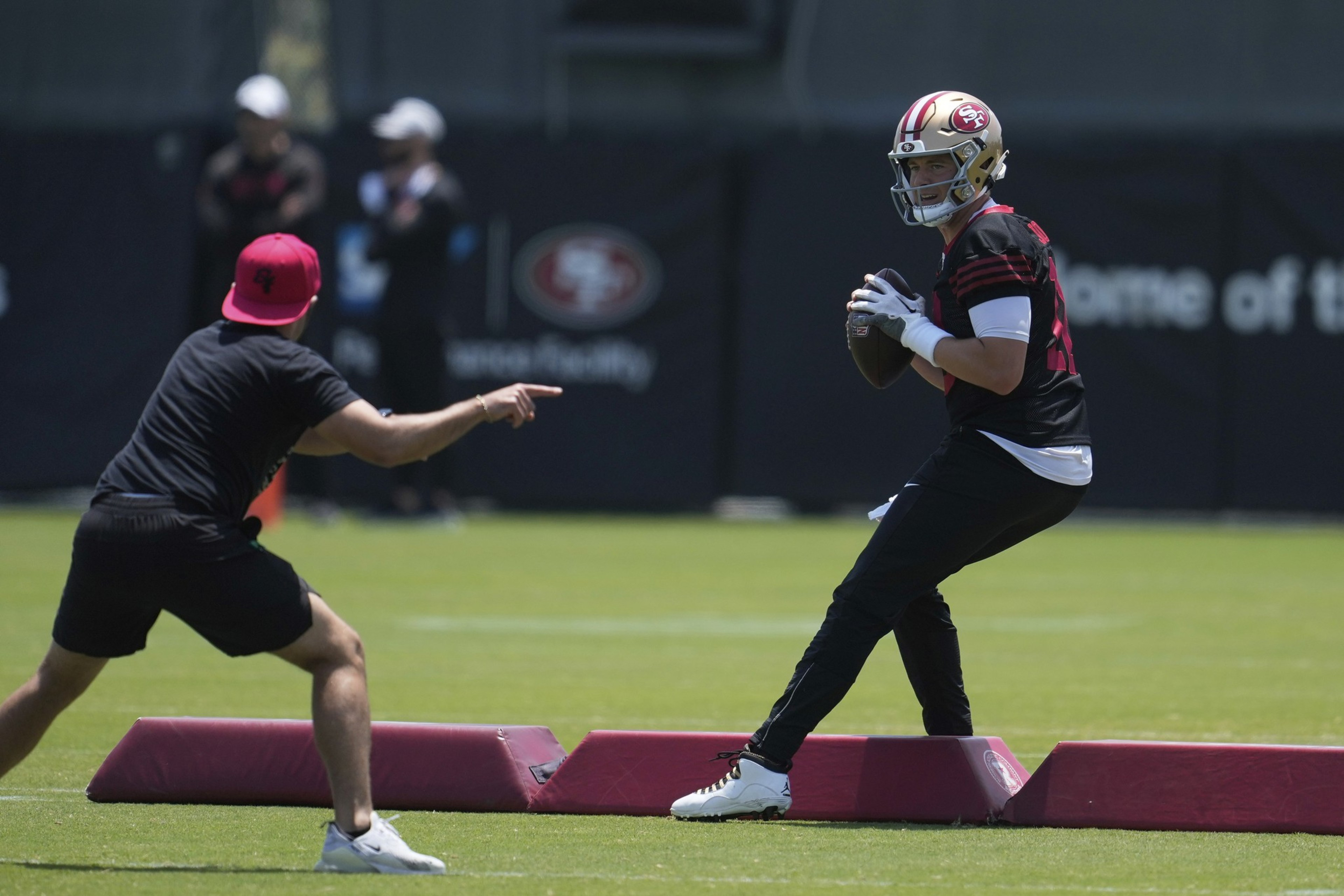 A football player in a helmet holds a ball, poised to throw. Another person in a red cap points at him during a practice session on a field.